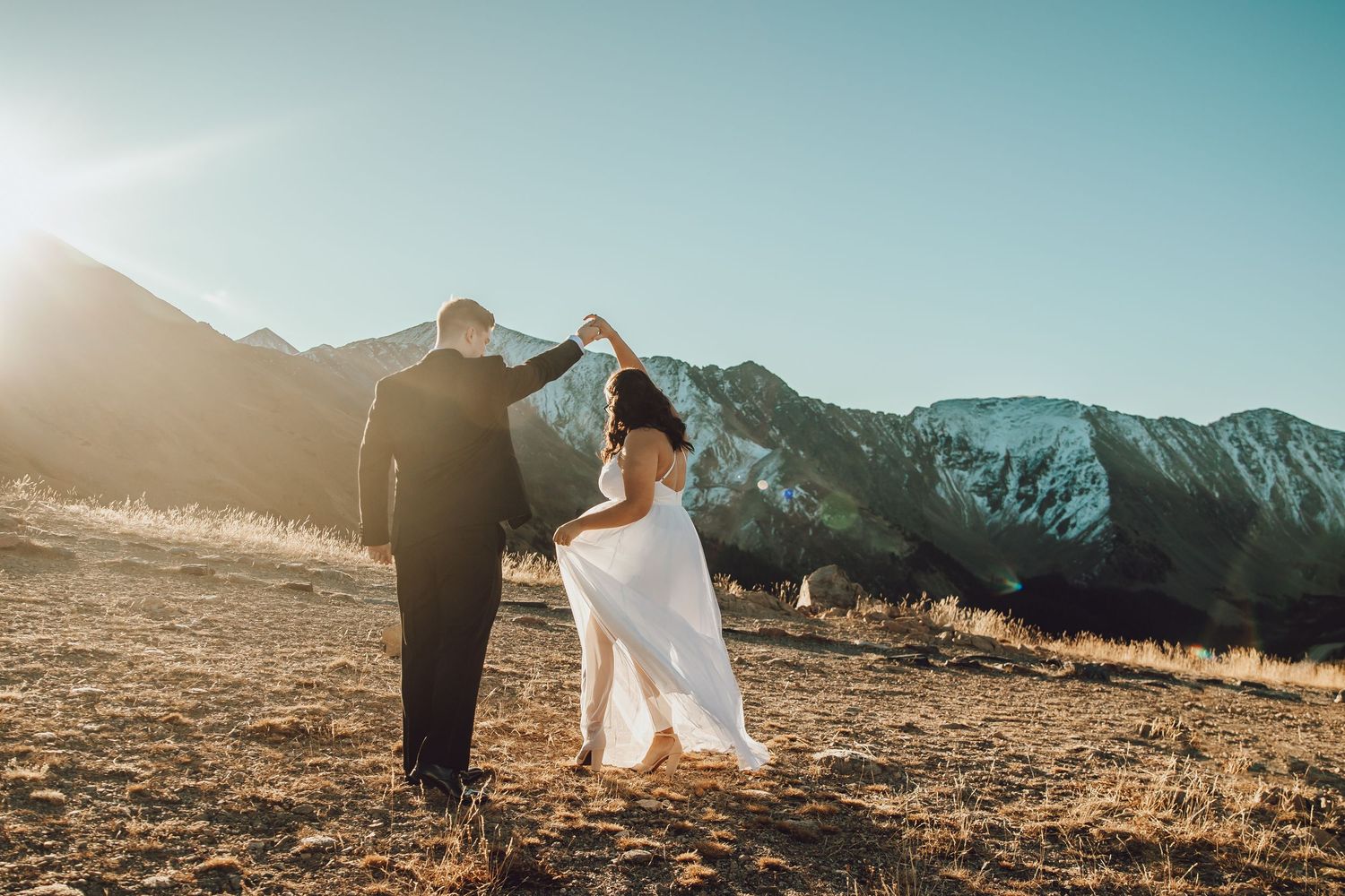 A couple in formal attire dances together on a mountain summit during golden hour with majestic peaks in the background.