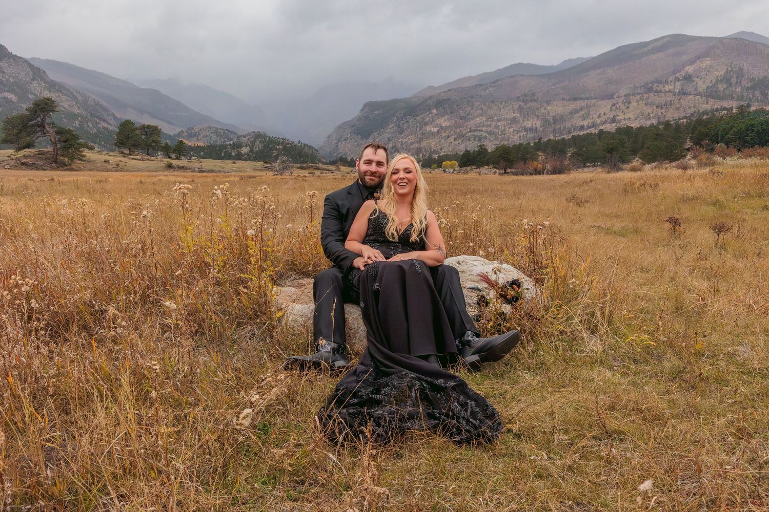 A couple sits together on a rock in a golden meadow with misty mountains in the backdrop in Scotland.