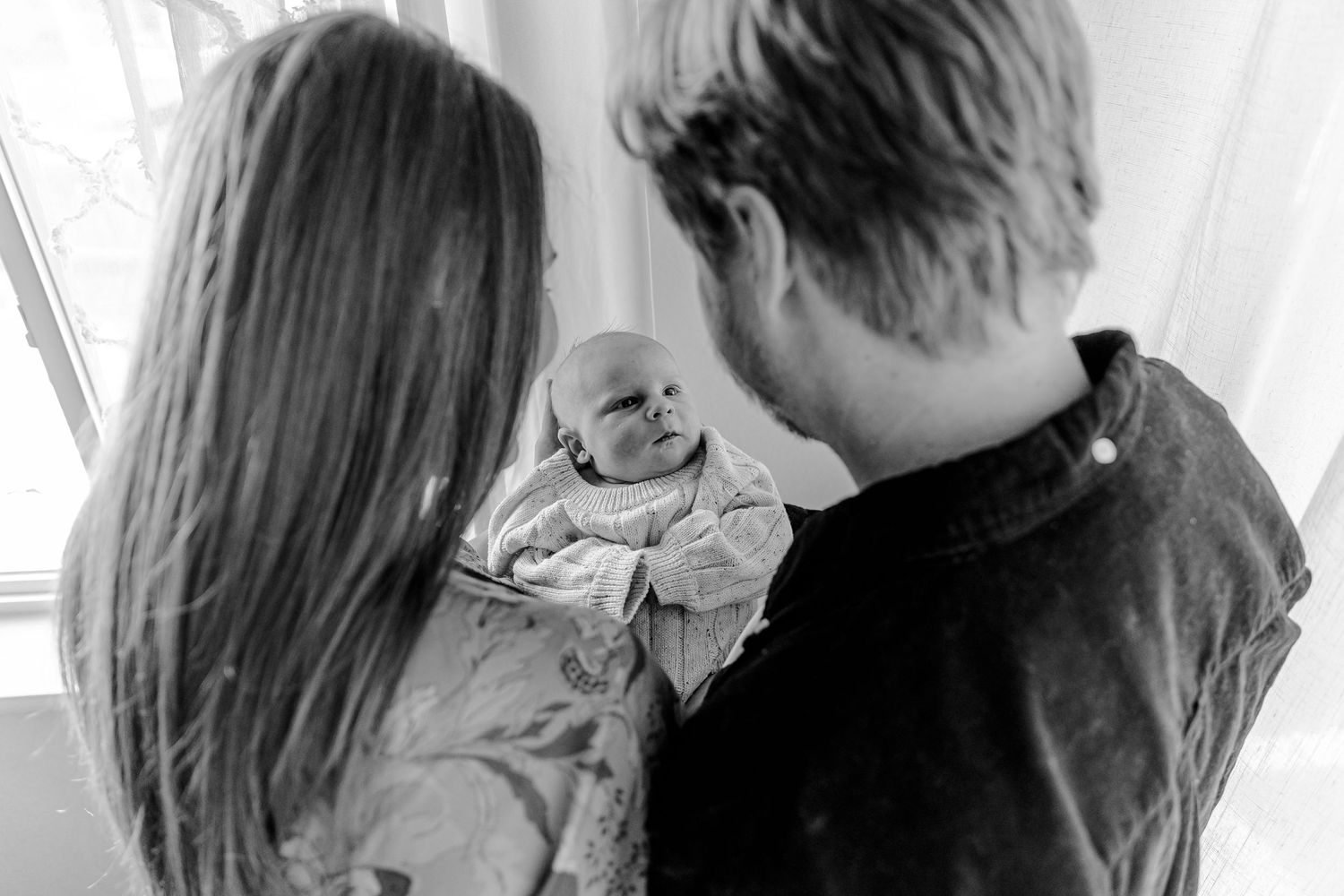 A couple shares a tender kiss in black and white while standing in front of a bright window.