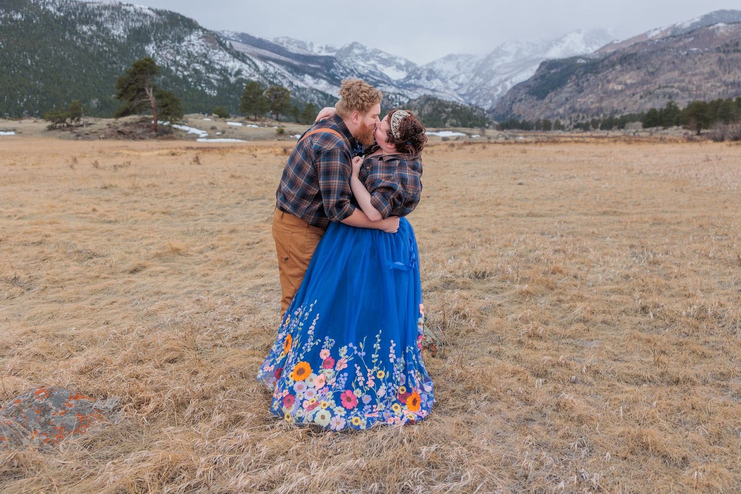 A couple embraces in a field with snow-capped mountains in the background, the woman wearing a vibrant blue floral dress.