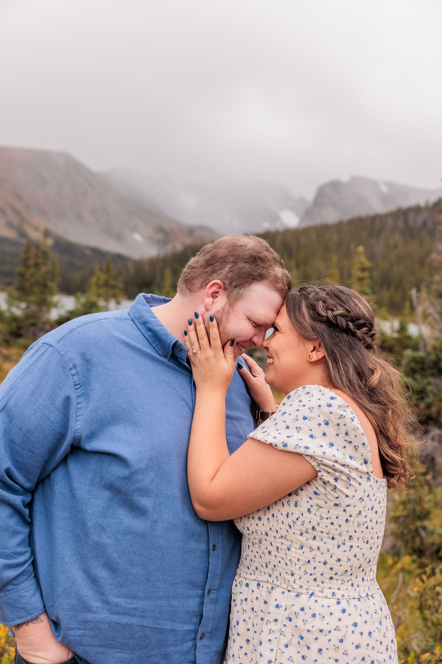 A romantic couple shares an intimate moment in front of misty mountain peaks and evergreen forest.