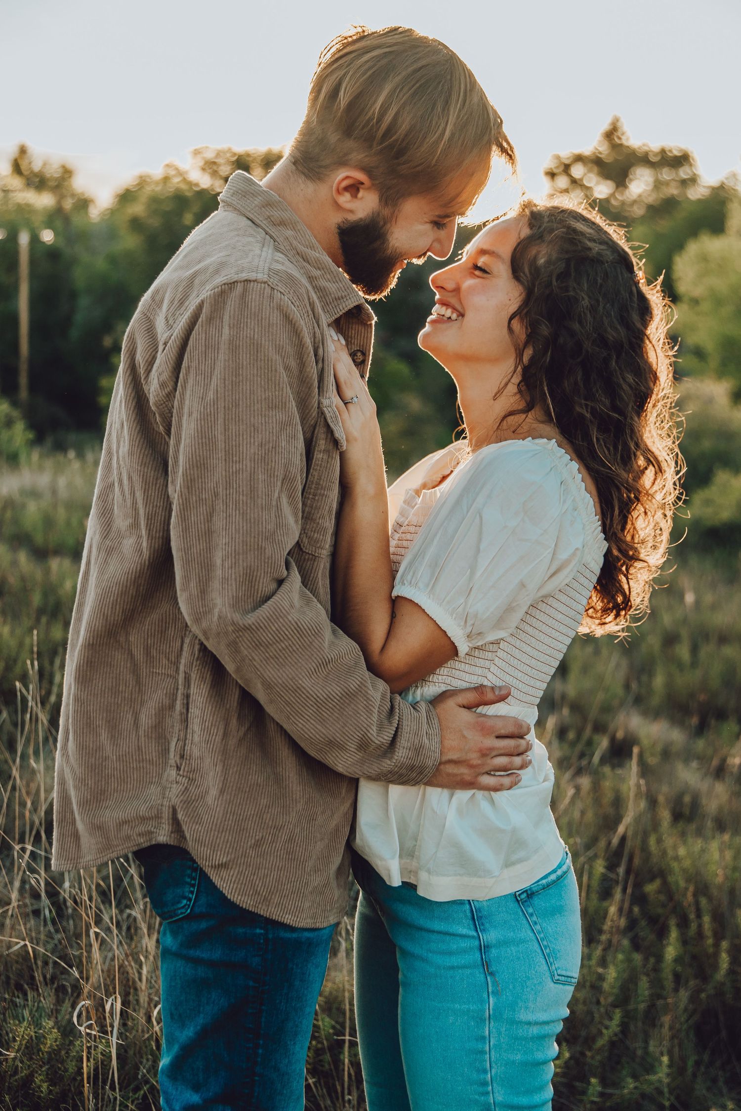 A romantic couple embraces in a golden sunlit field wearing blue jeans and neutral tops during a sunset photoshoot.