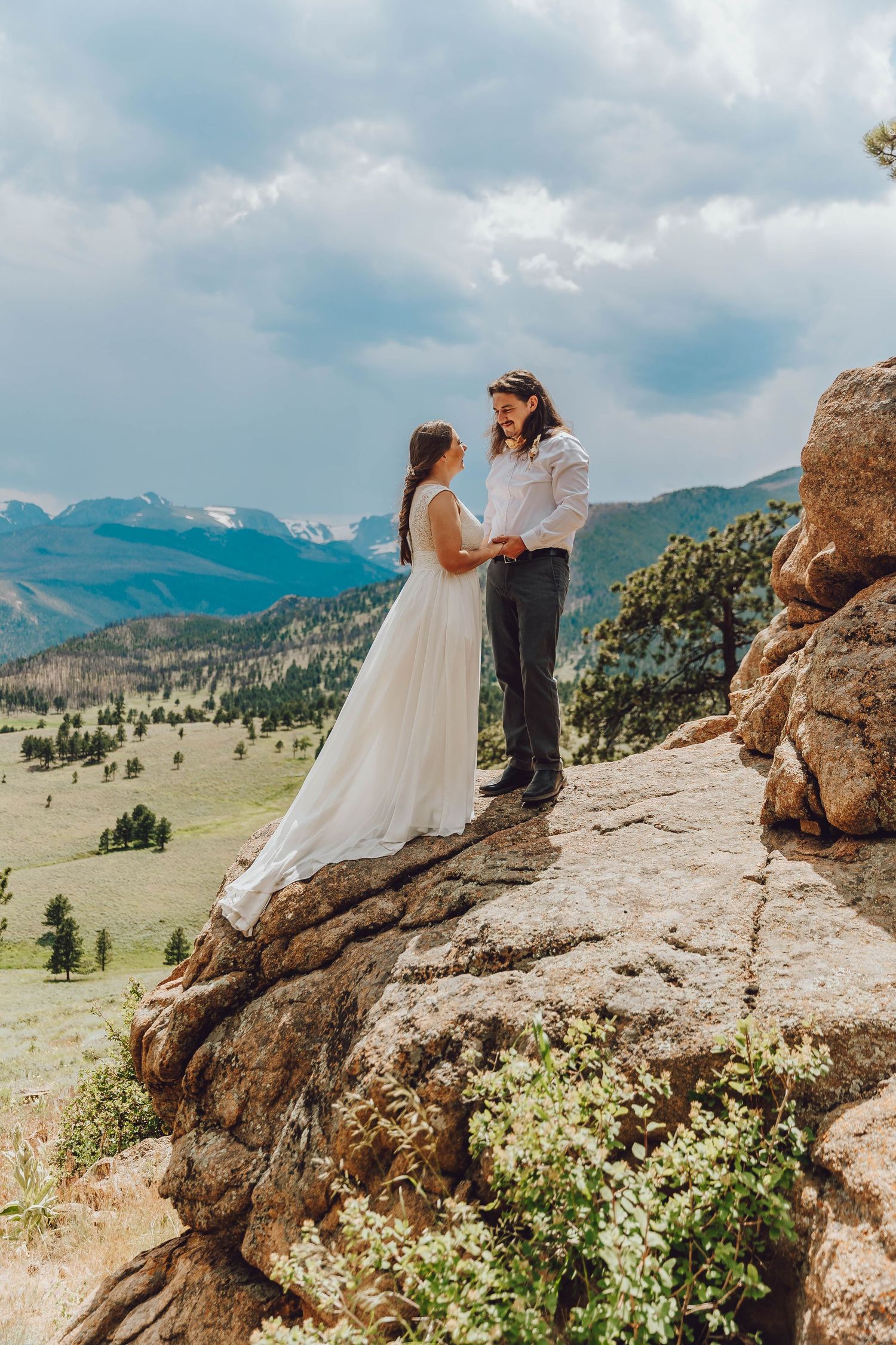 A wedding couple stands on a rocky mountain overlook with sweeping mountain vistas and blue skies in the background.