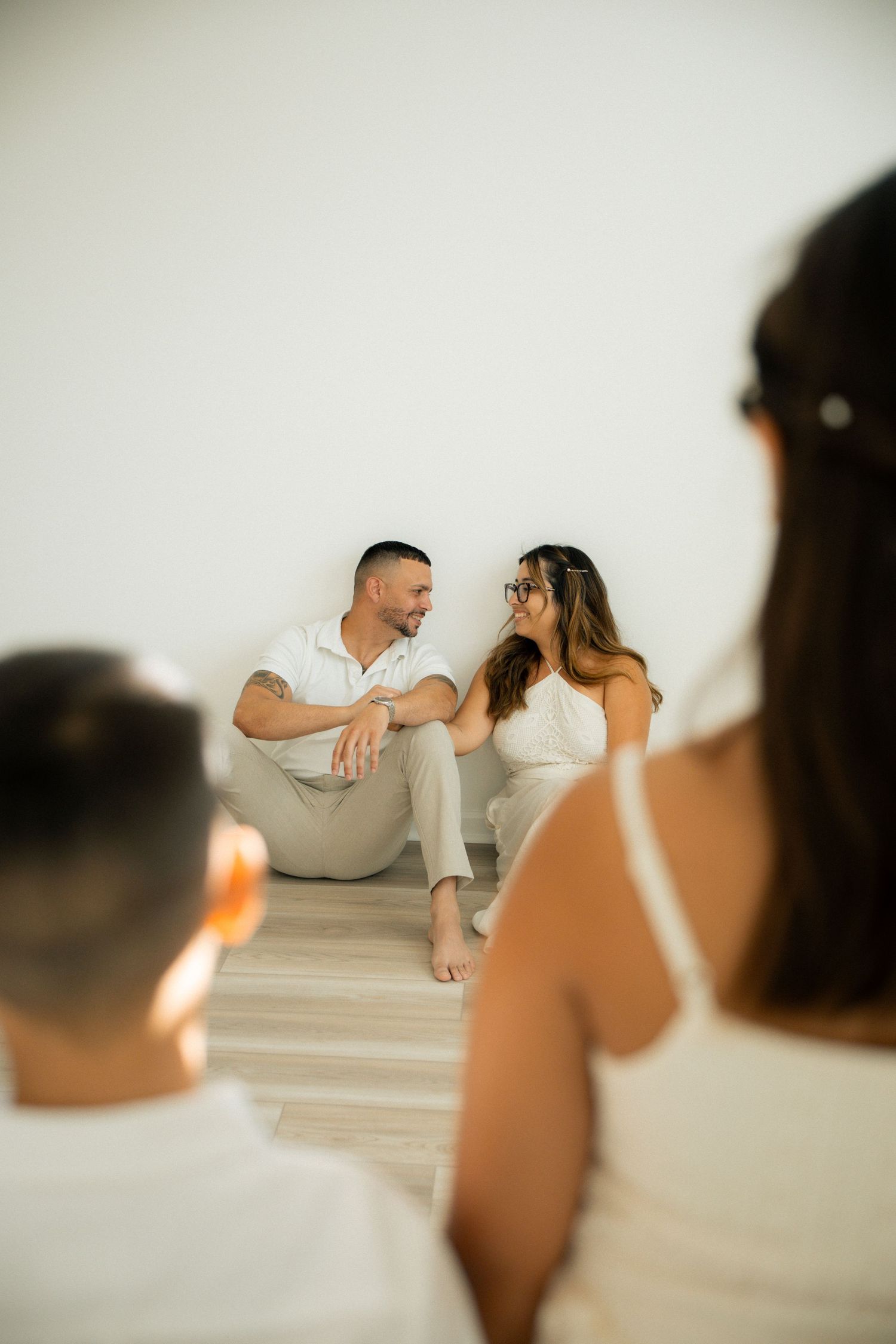 Couple in white outfits sitting on floor gazing at each other during a romantic photo session.