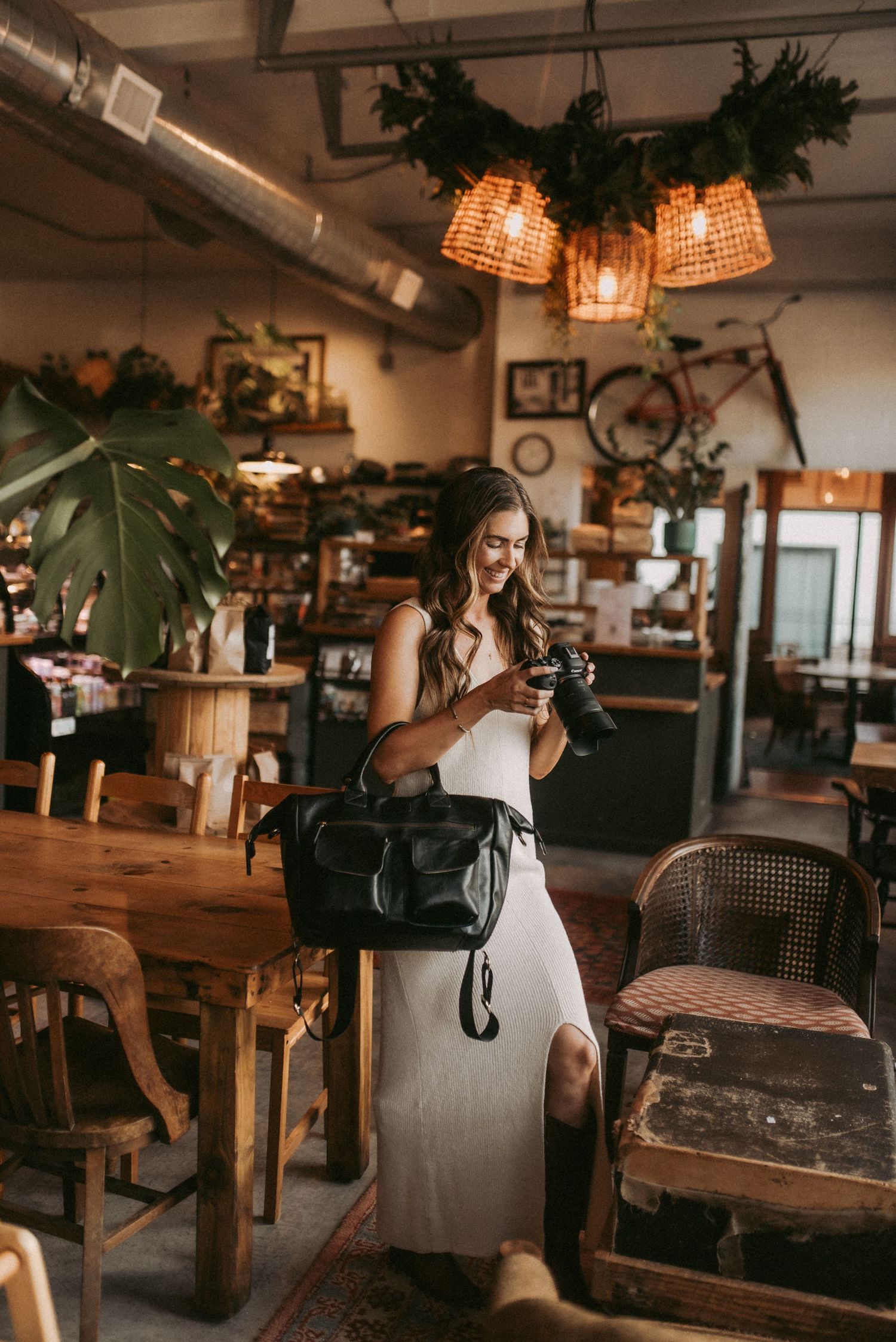 Woman in white dress holding camera and black bag stands in a rustic cafe with warm pendant lights and wooden furniture.