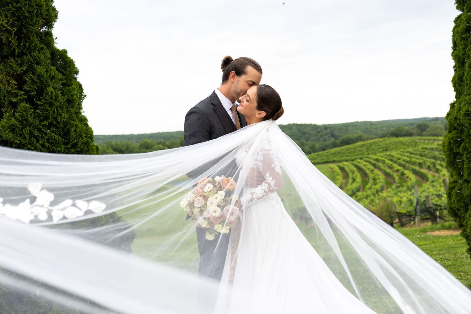 Bride and groom share a romantic kiss at a vineyard wedding, with her flowing veil billowing in the breeze.