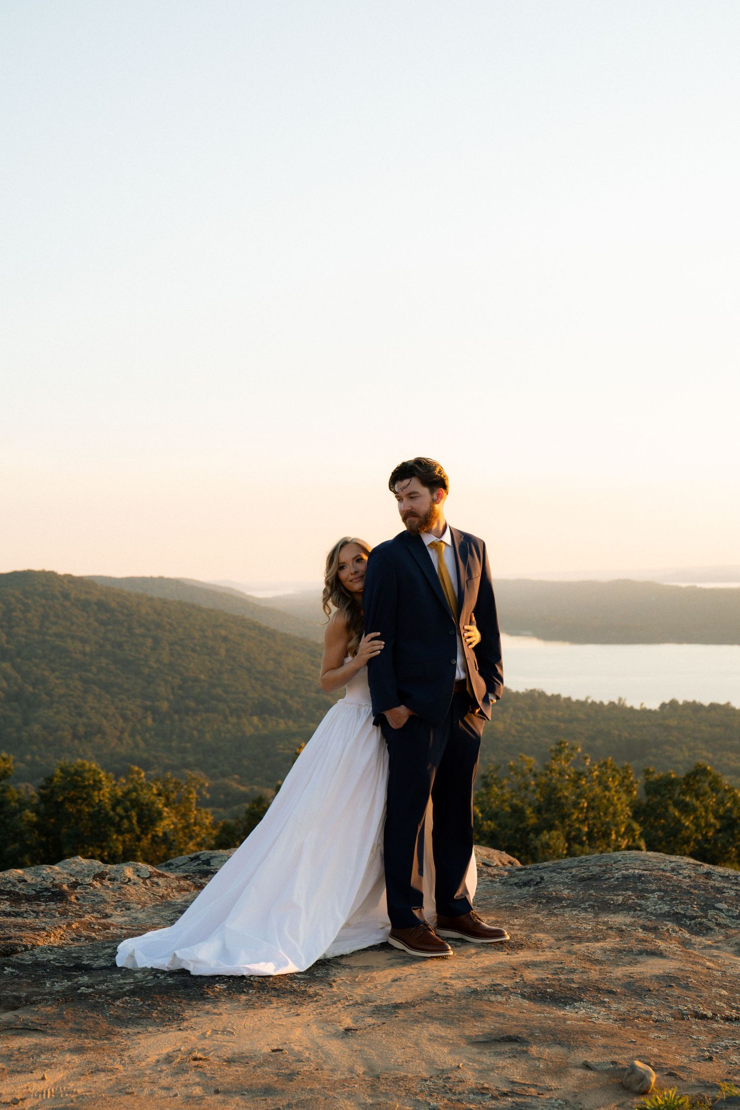 Bride and groom pose on a rocky mountaintop at sunset with scenic lake and forest views below.