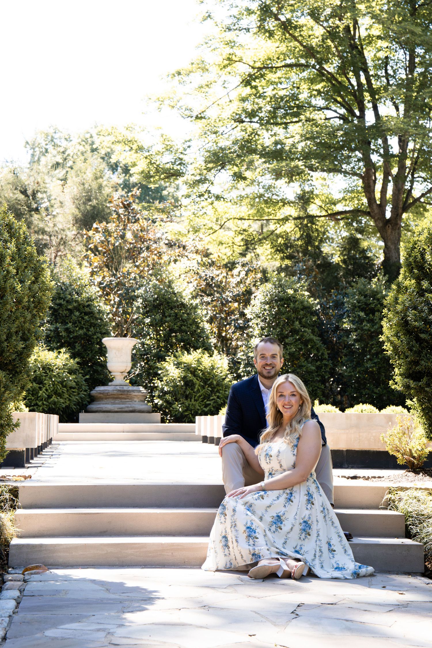 Couple posing on garden steps surrounded by lush greenery, woman in floral dress and man in navy suit.