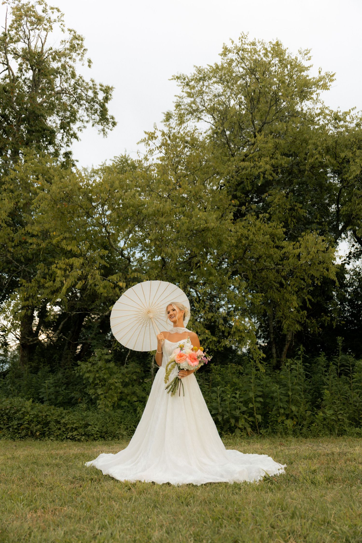 Bride in white gown holding floral bouquet and parasol umbrella standing outdoors with lush green trees background.