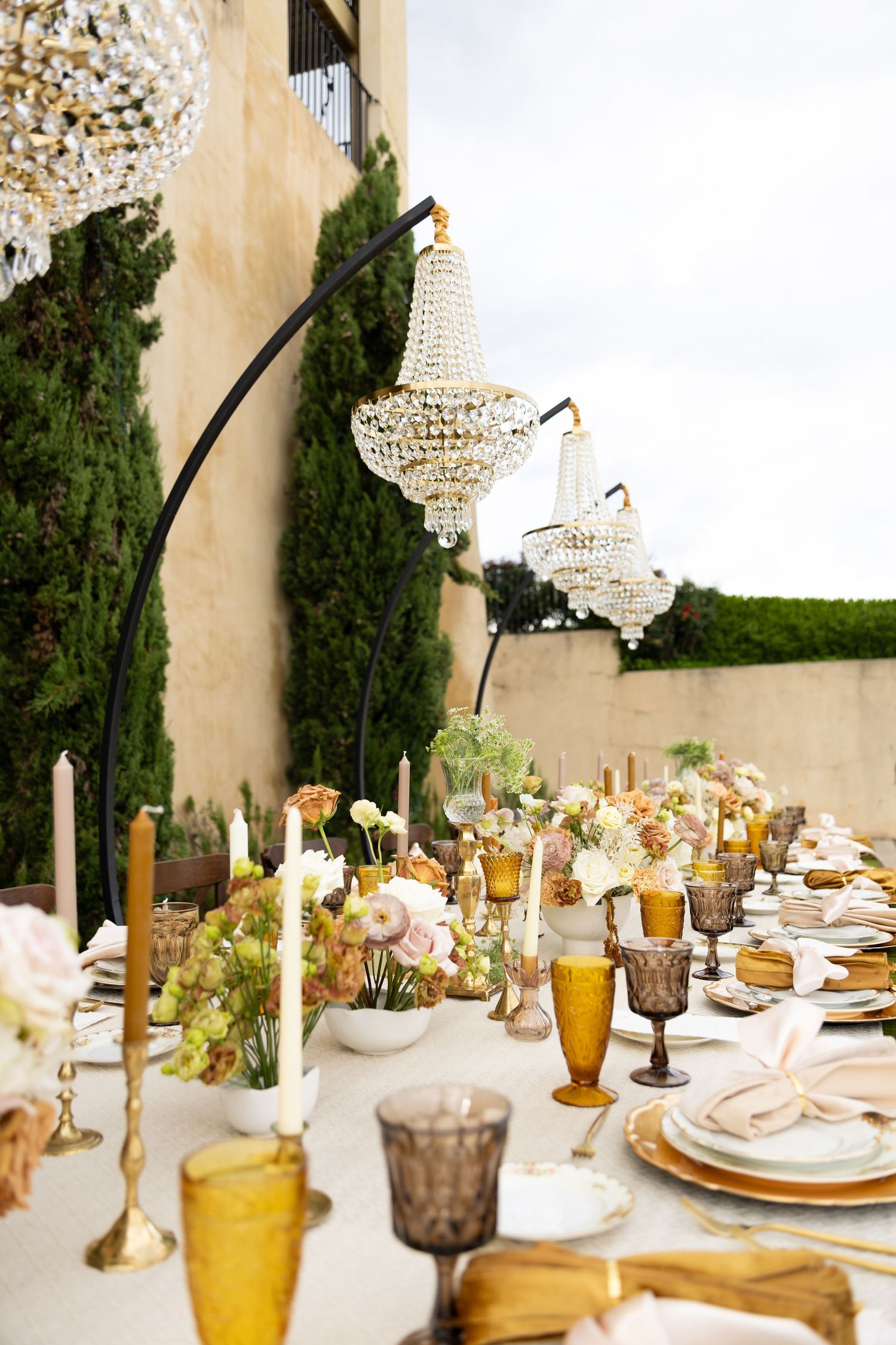 Elegant outdoor dining table with crystal chandeliers, amber glassware, floral centerpieces, and gold accents for a luxury event.