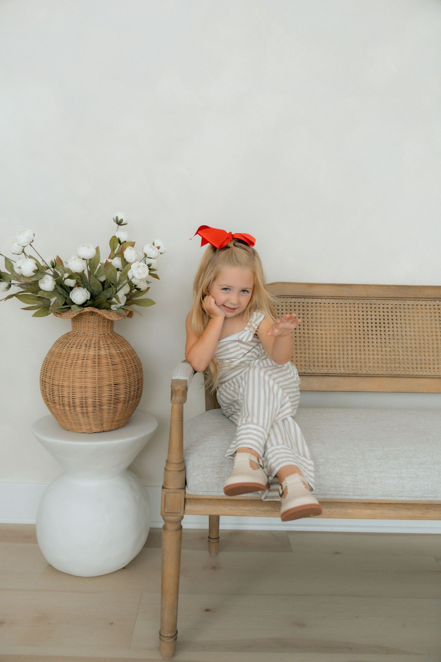 Young girl with red bow sits smiling on wooden bench beside wicker vase with white flowers.