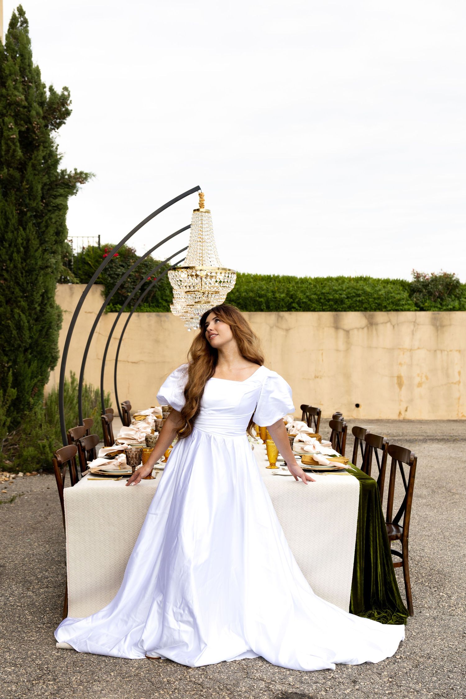 Bride in white puff-sleeve gown stands by elegant outdoor dining table with chandelier and greenery backdrop.