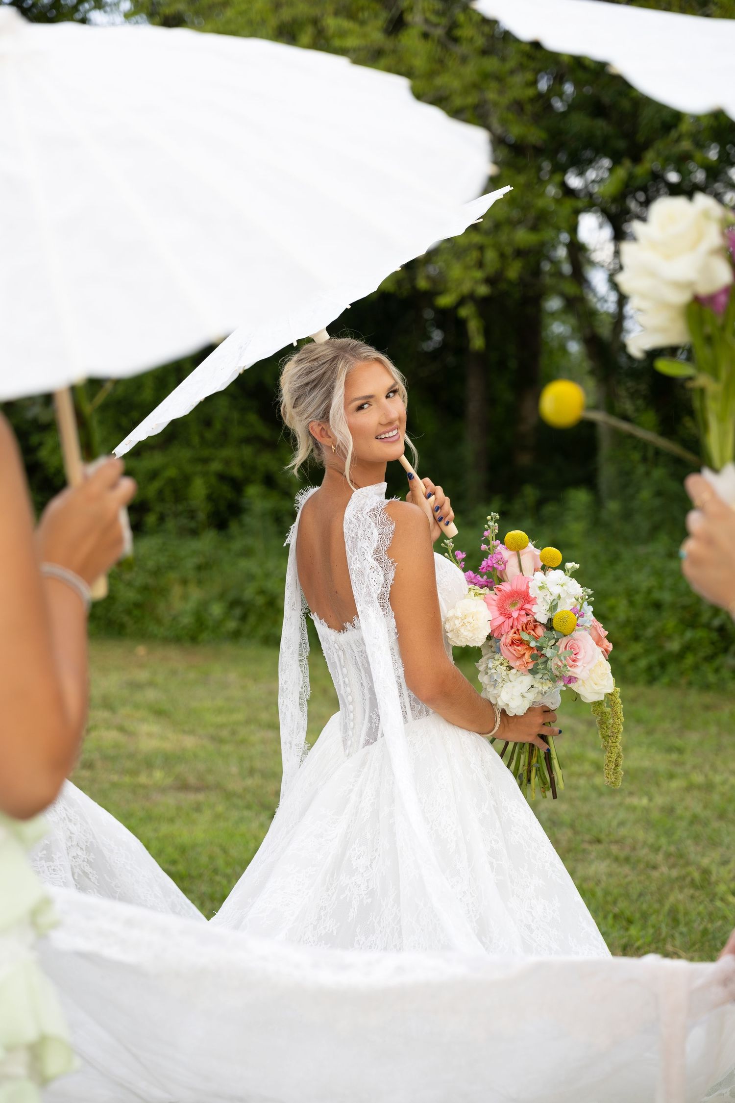 Bride in backless lace gown holds colorful bouquet outdoors, surrounded by guests with white parasols at garden wedding.