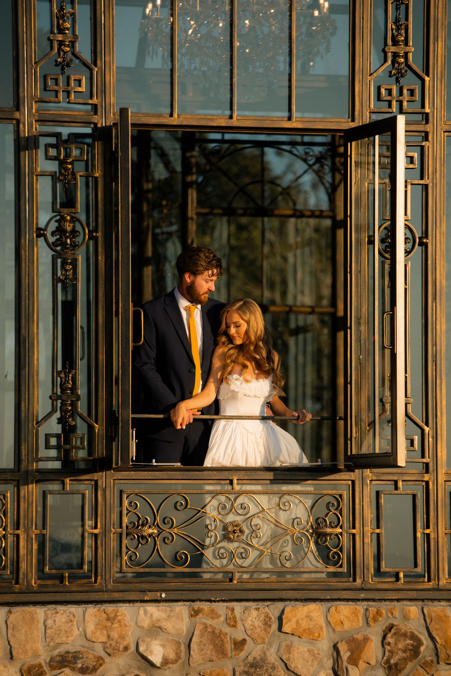 Bride and groom share a romantic moment in an ornate golden wrought iron window at sunset.