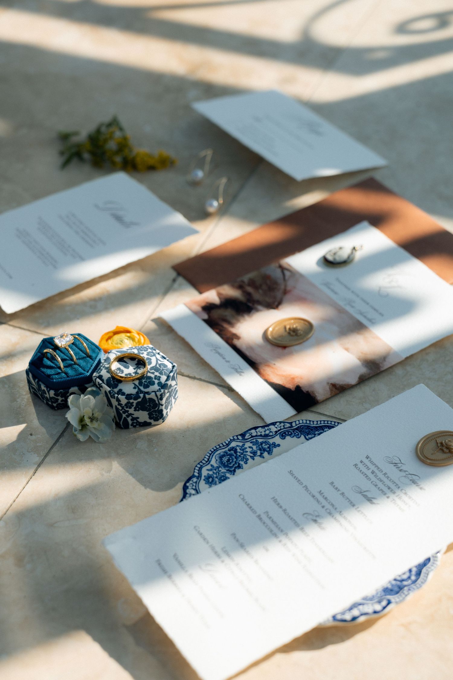 Elegant wedding stationery suite with blue and white ceramic ring box, gold wax seals, and floral accents on a sunlit surface.