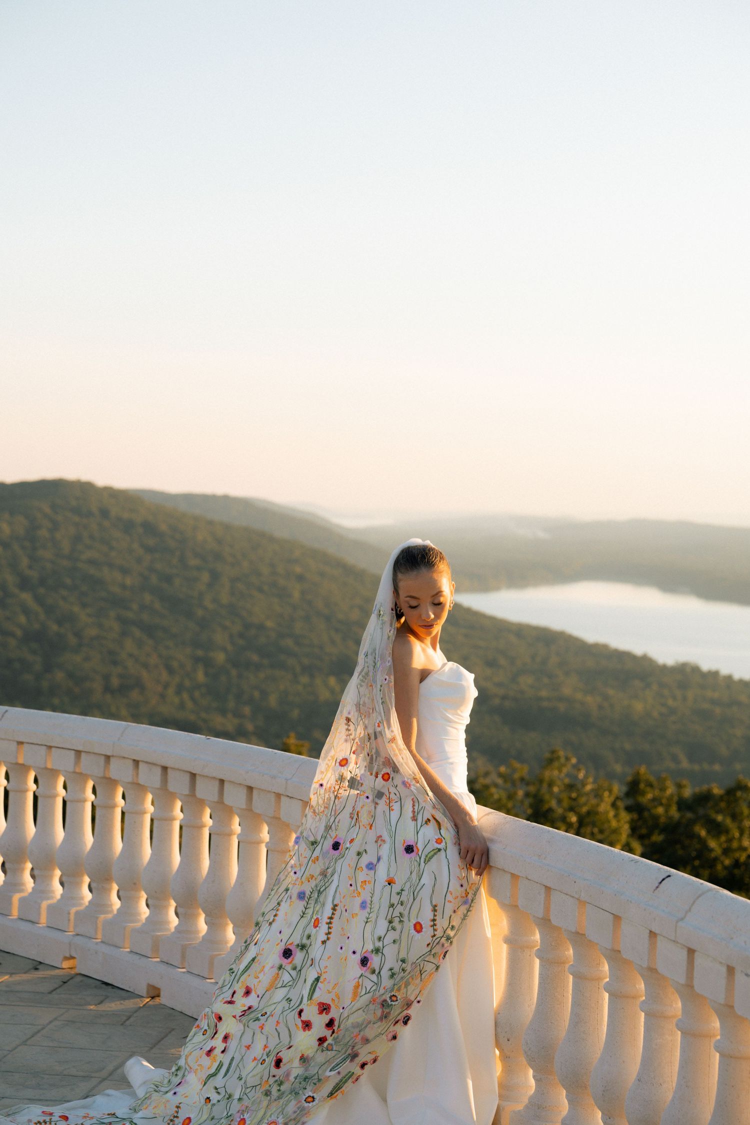 Bride in white gown with floral veil poses on balcony overlooking lush green hills and lake at golden hour.