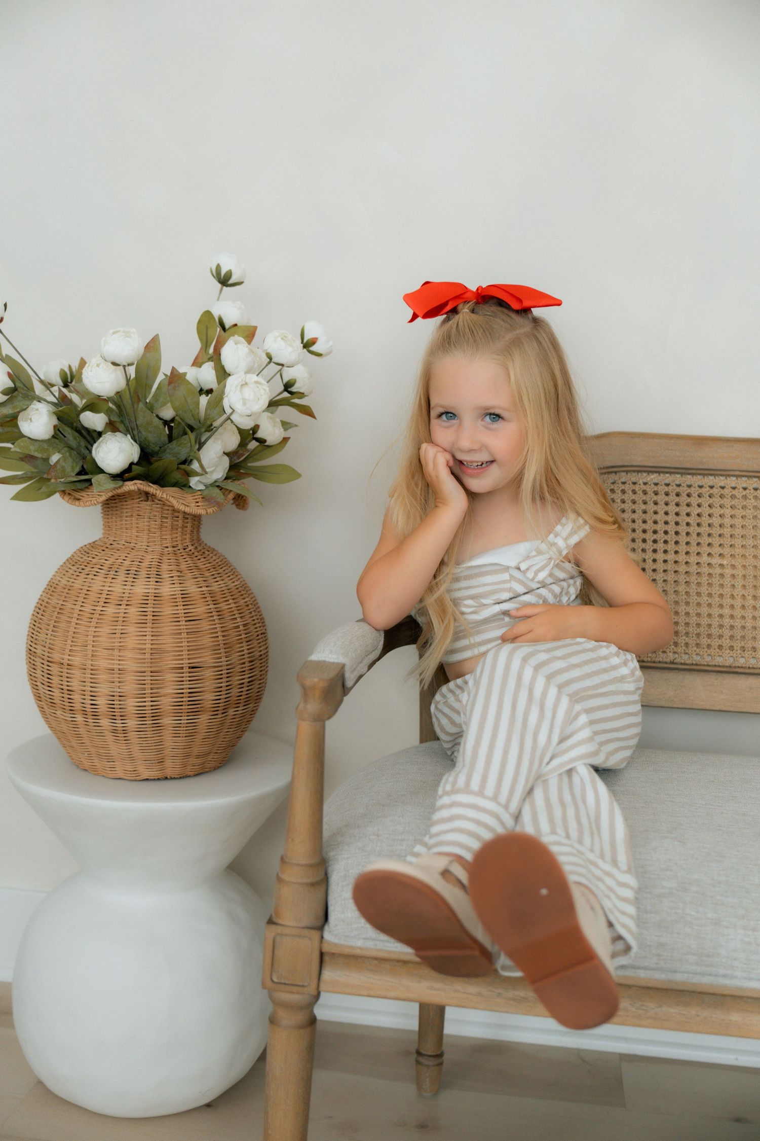 Young blonde girl in striped jumpsuit with red bow sits on wooden bench beside wicker vase with white flowers.