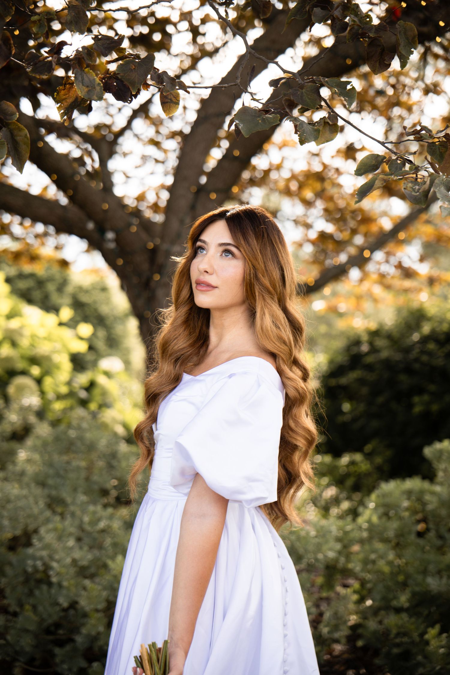 Young woman in white off-shoulder dress with long wavy hair standing under a tree in a lush garden.