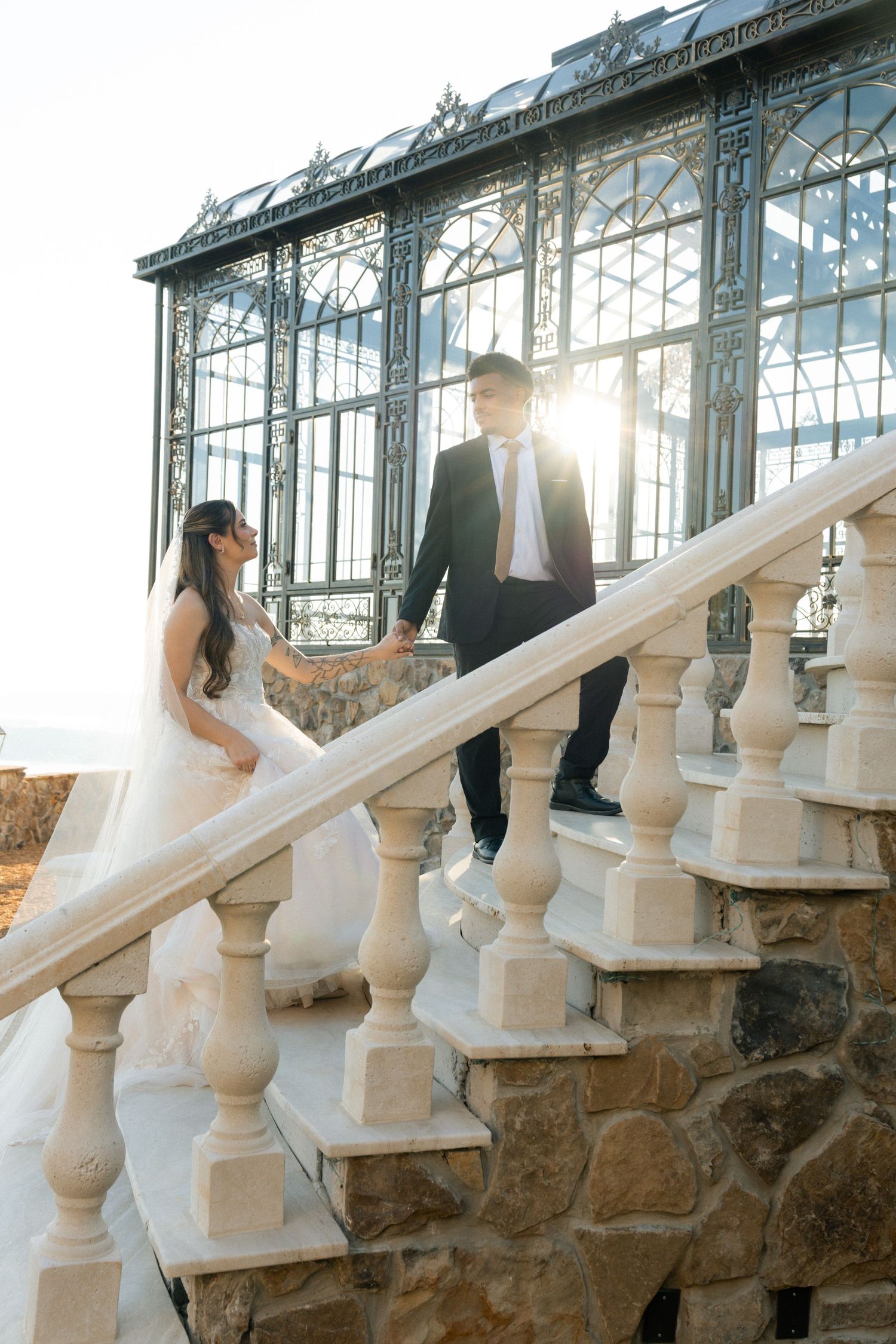 Bride and groom holding hands on elegant white stone staircase outside ornate glass greenhouse venue.