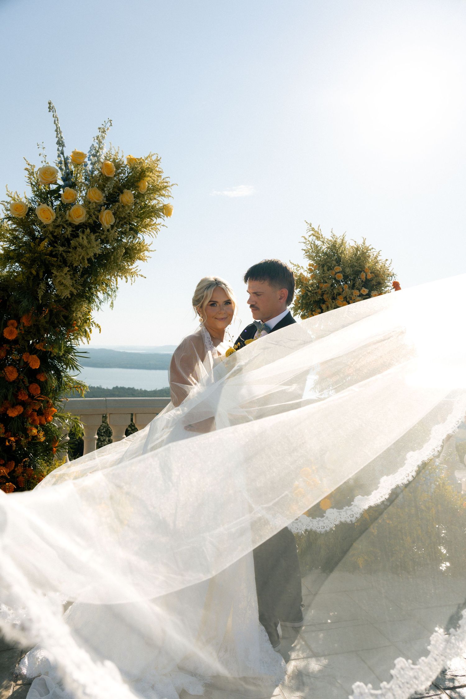 Bride and groom embrace at outdoor wedding ceremony with yellow floral arch, veil flowing in wind against sunny sky.