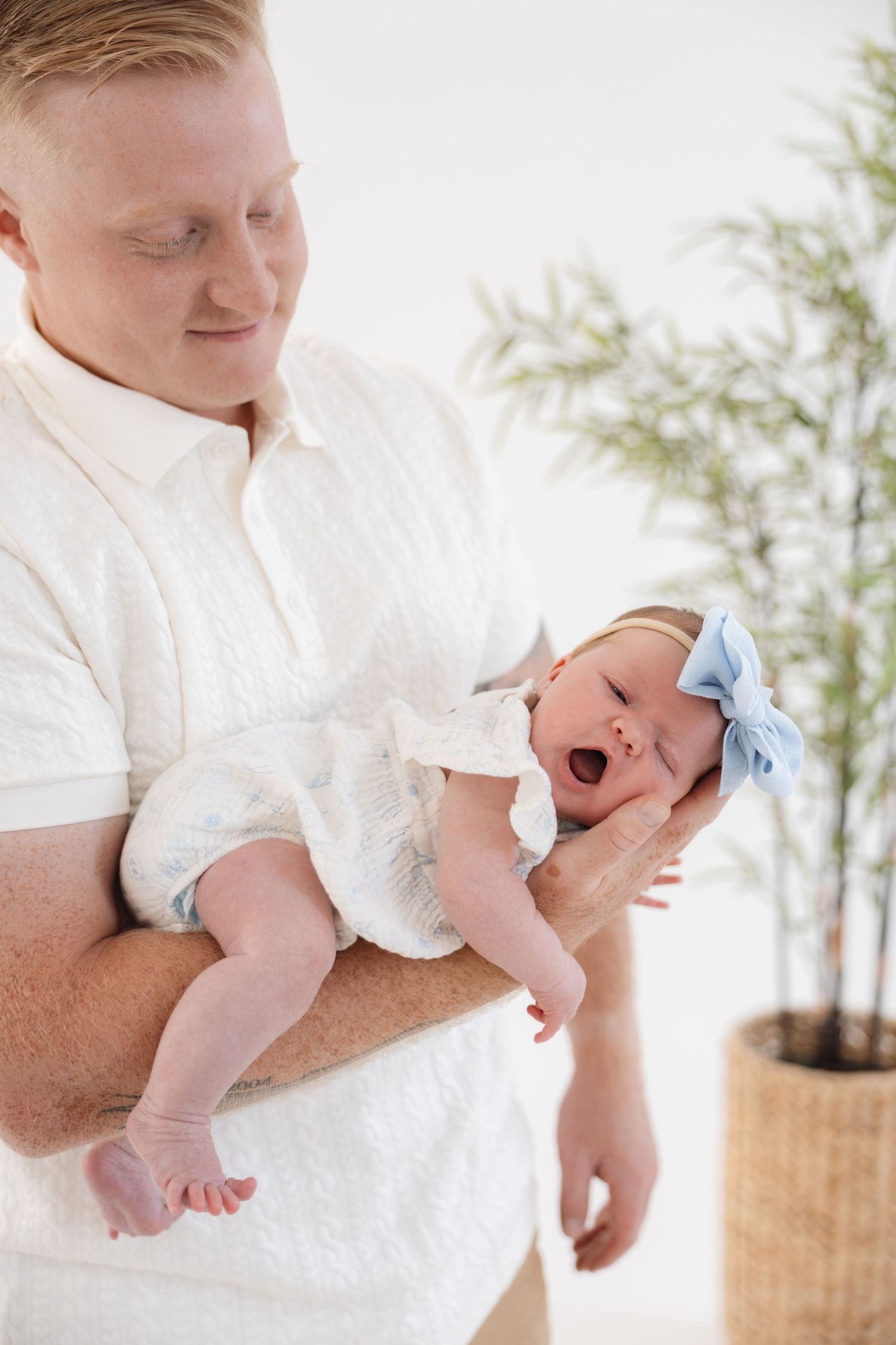 A father in a white shirt gently holds his yawning newborn baby girl wearing a blue bow headband.