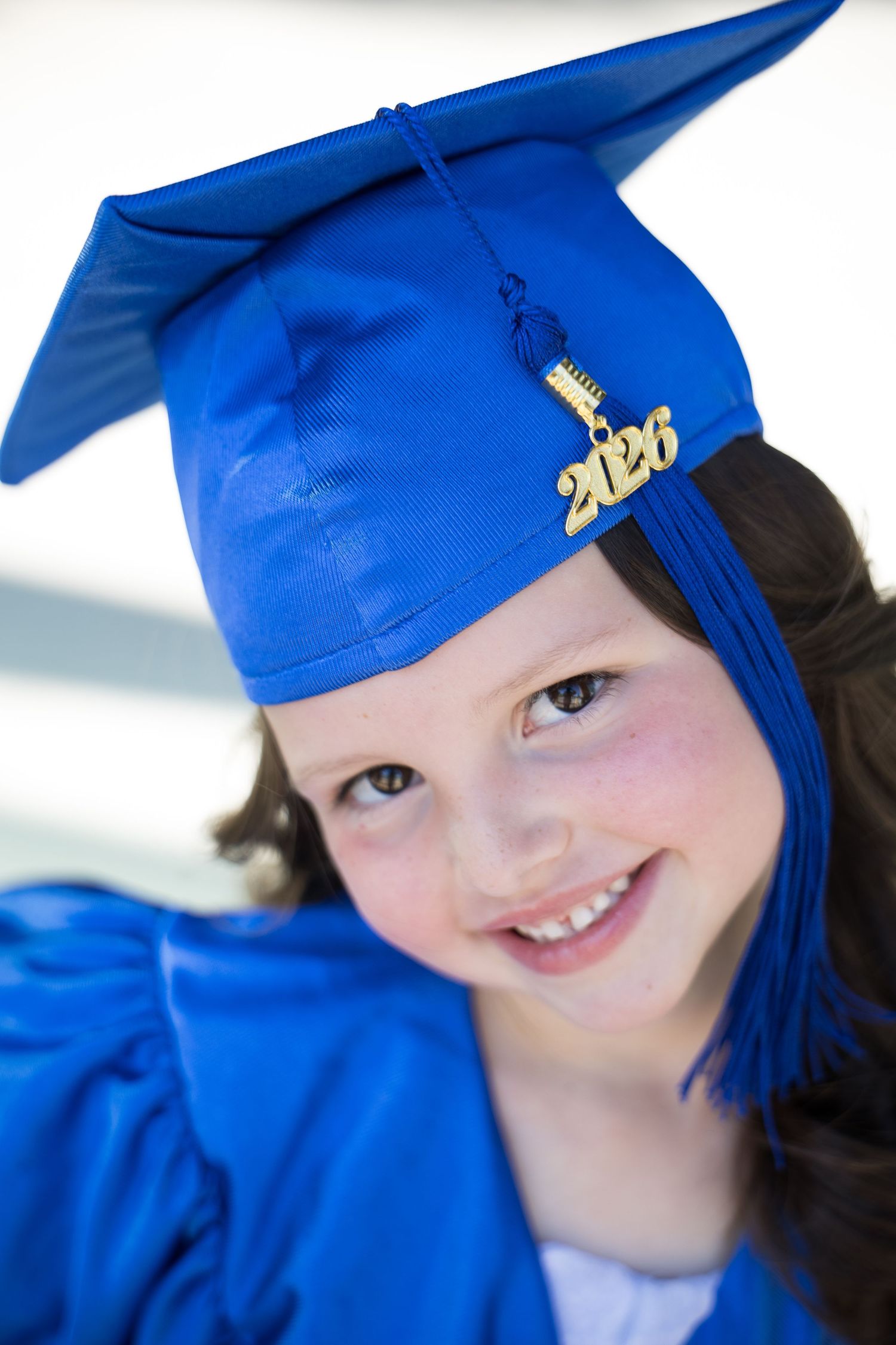 A young girl smiles wearing a blue graduation cap and gown with a 2026 tassel for her kindergarten ceremony.