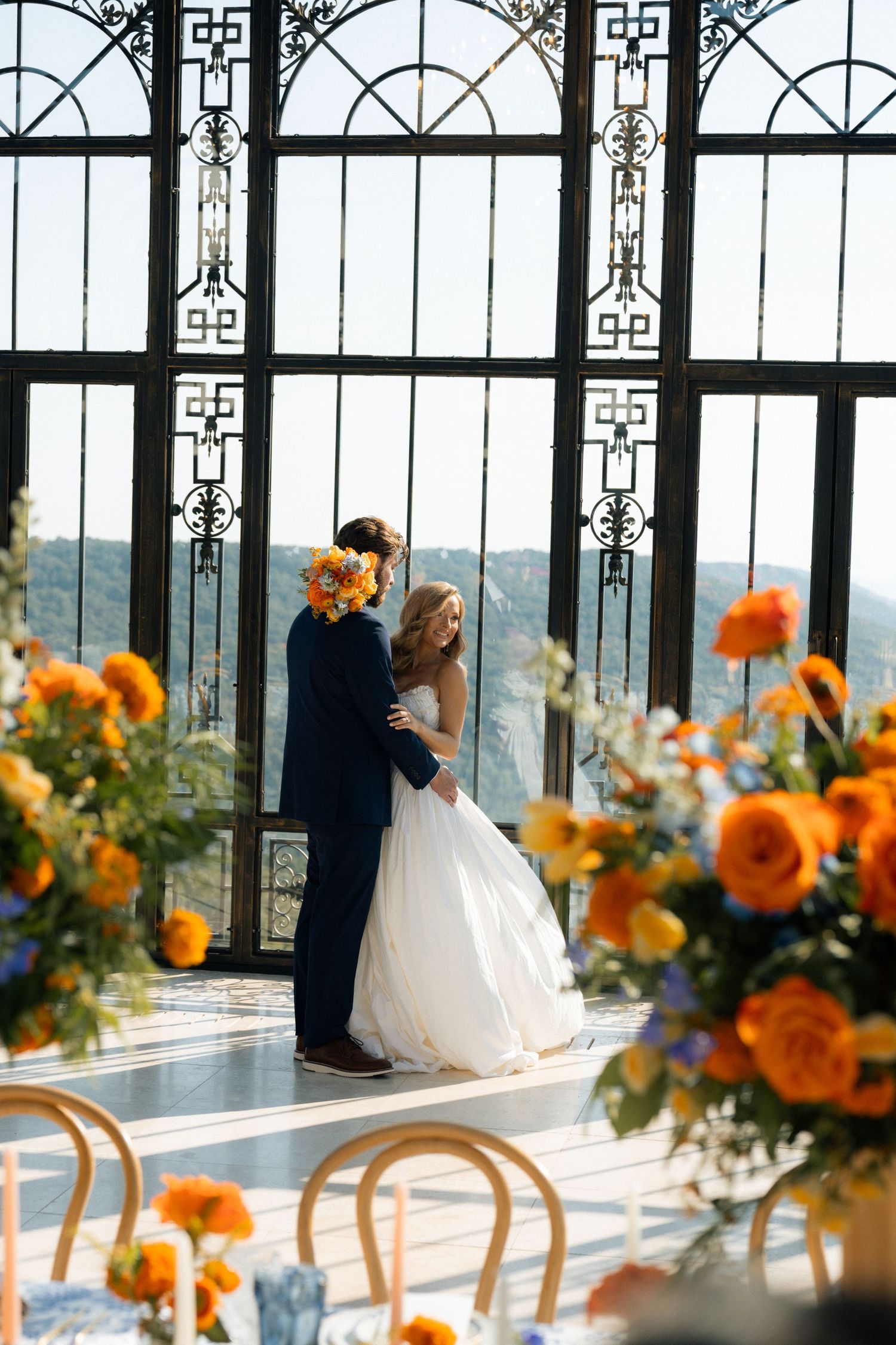 Bride and groom share a romantic moment at an elegant indoor wedding ceremony with orange floral arrangements.
