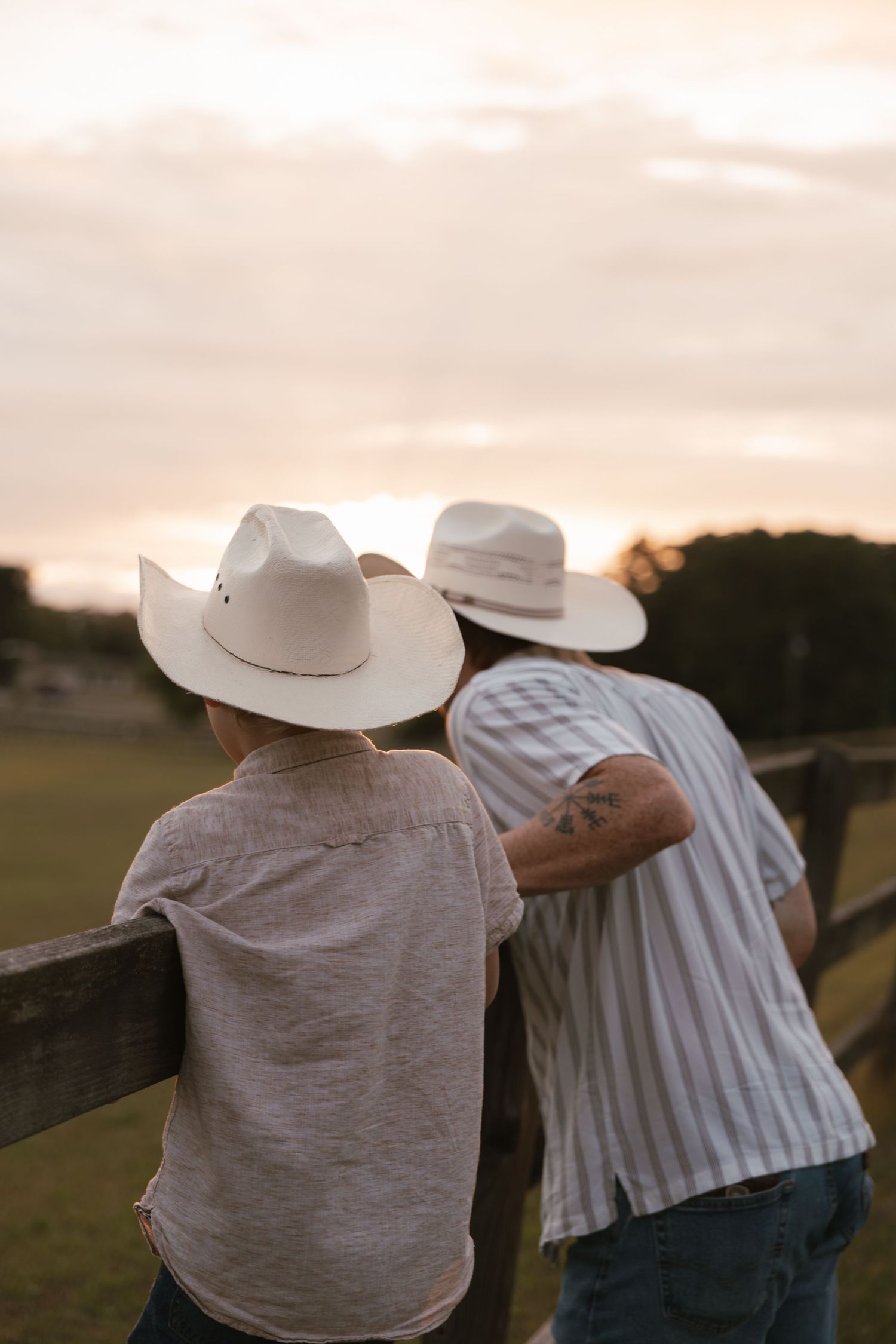 Two people in cowboy hats lean on a wooden fence at sunset on a rural farm.