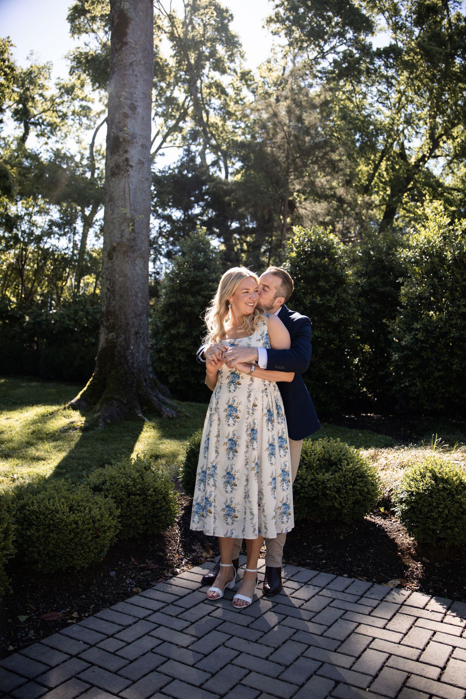Couple embracing on brick path in lush garden, woman in floral dress, man in navy suit, surrounded by greenery.