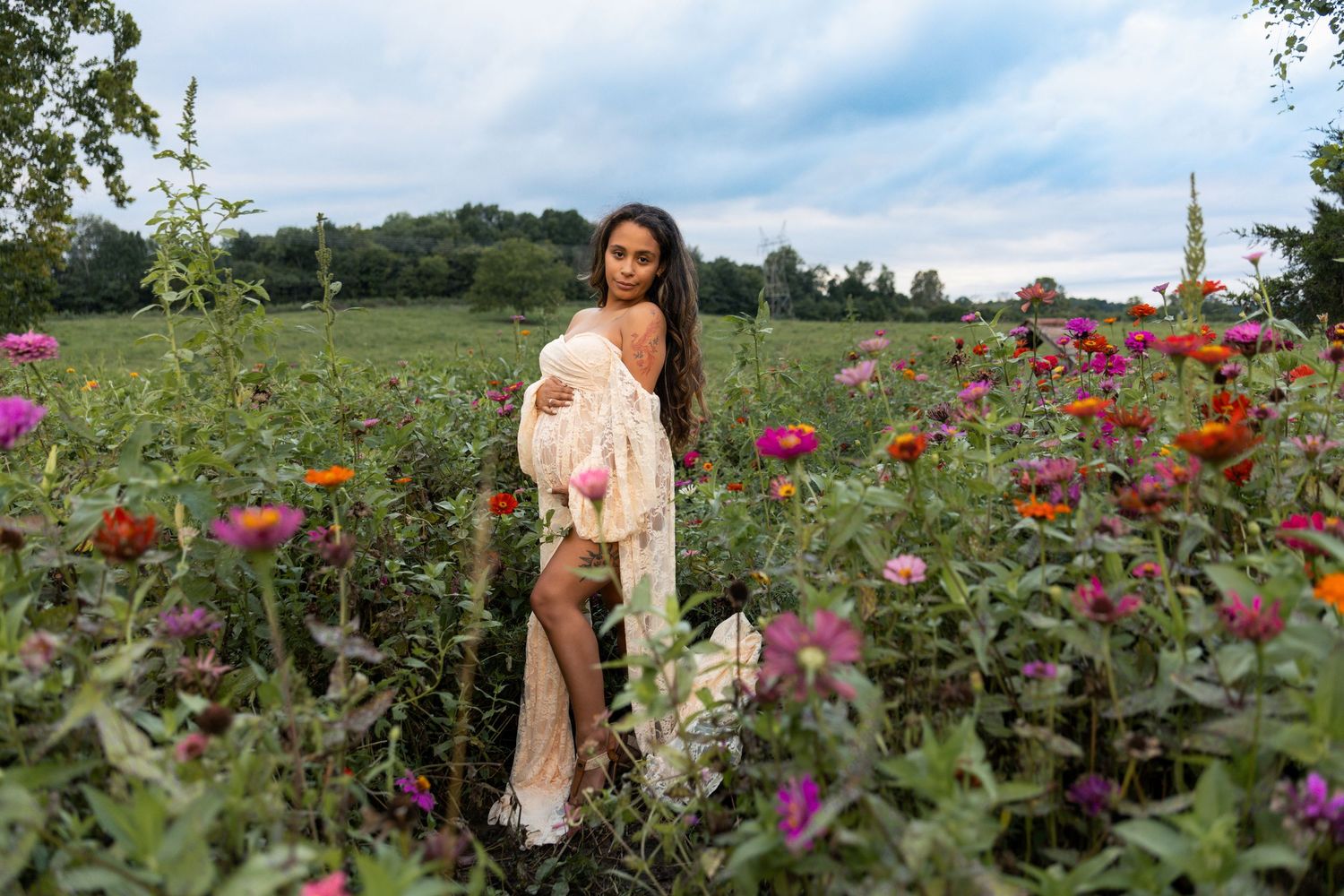 Woman in sheer white dress standing among colorful wildflowers in a lush green field under cloudy skies.