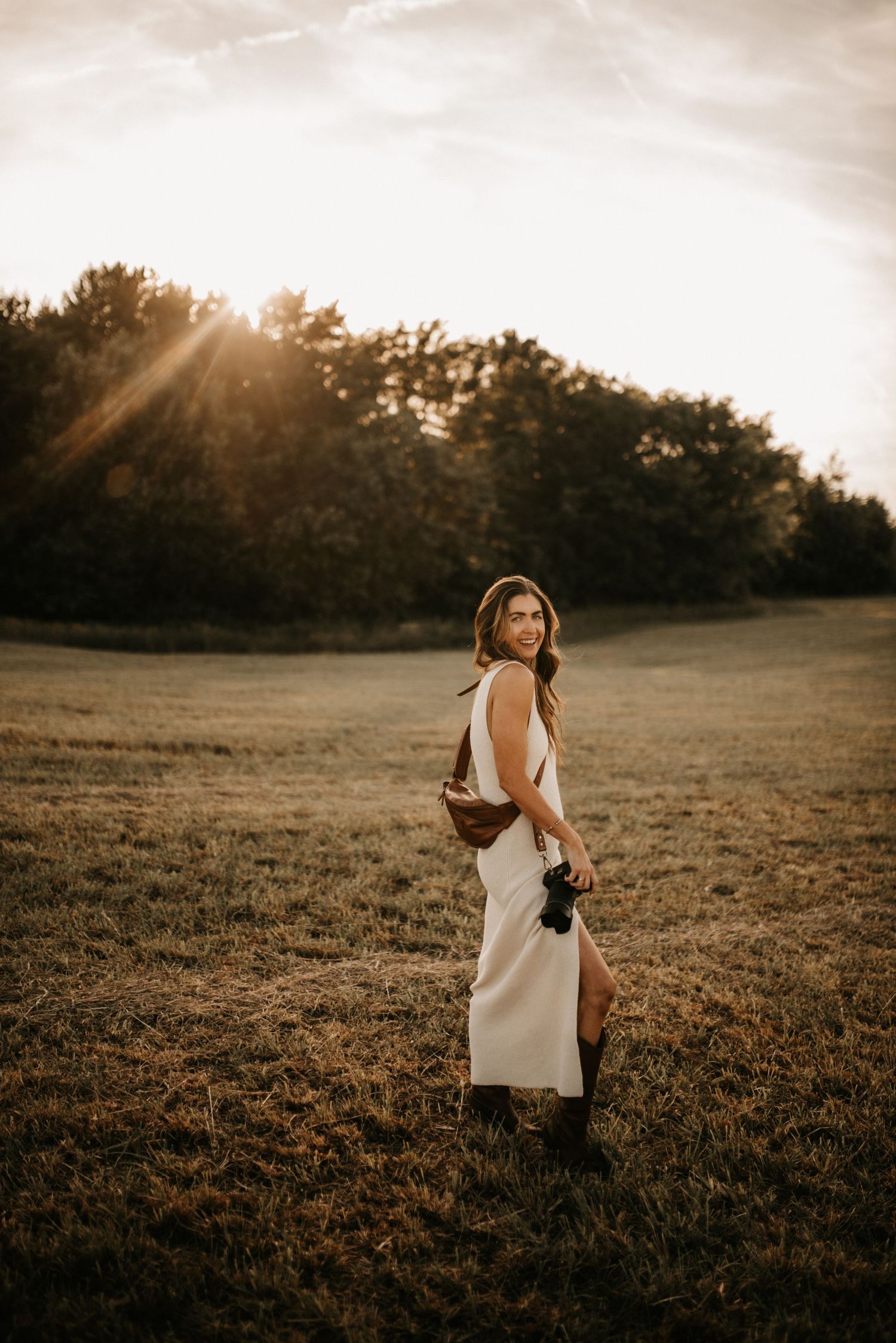 Woman in white dress and boots holds camera in golden hour field with sun rays and trees in background.
