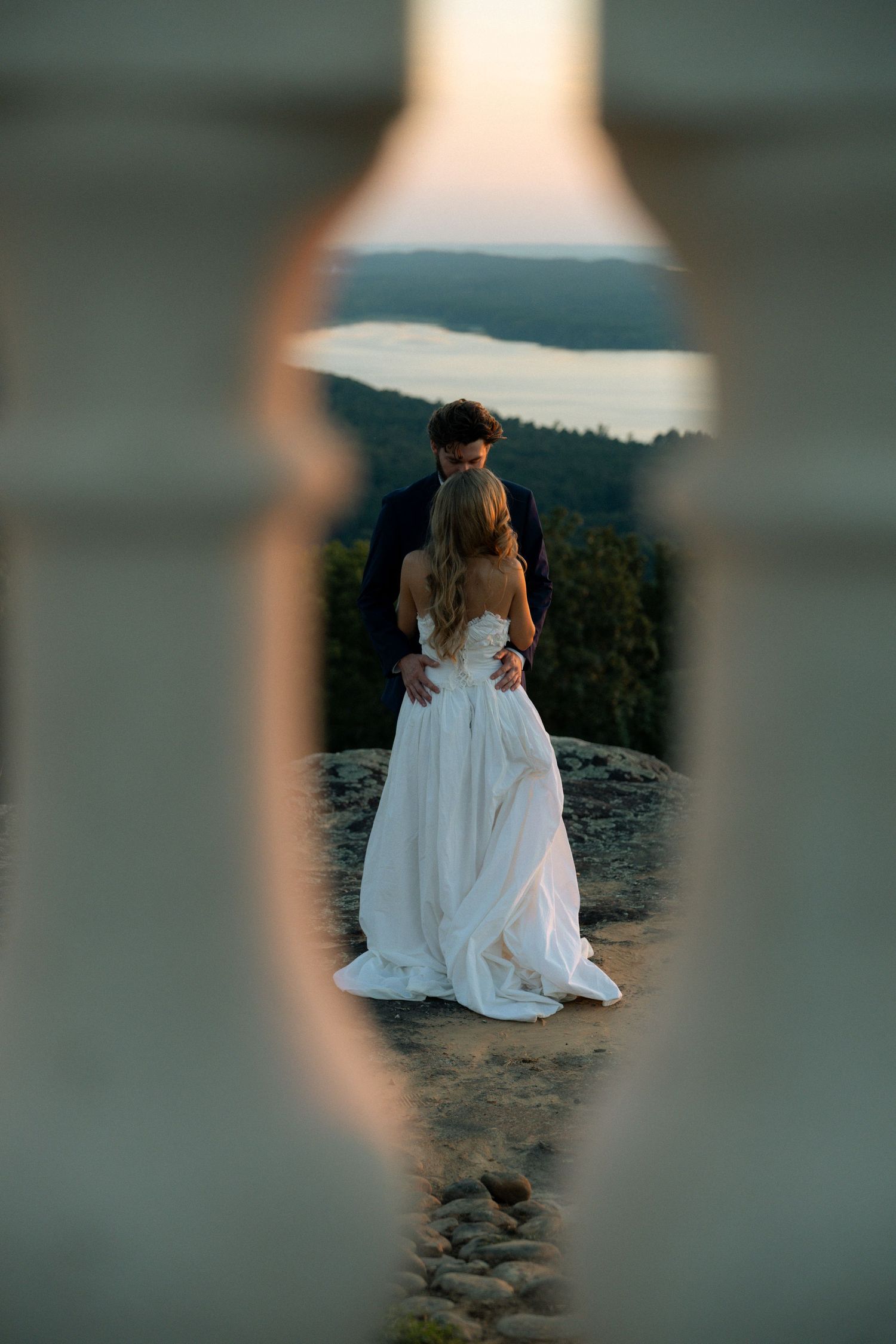 Bride and groom embrace on rocky overlook at sunset, framed through decorative white fence opening with lake view.