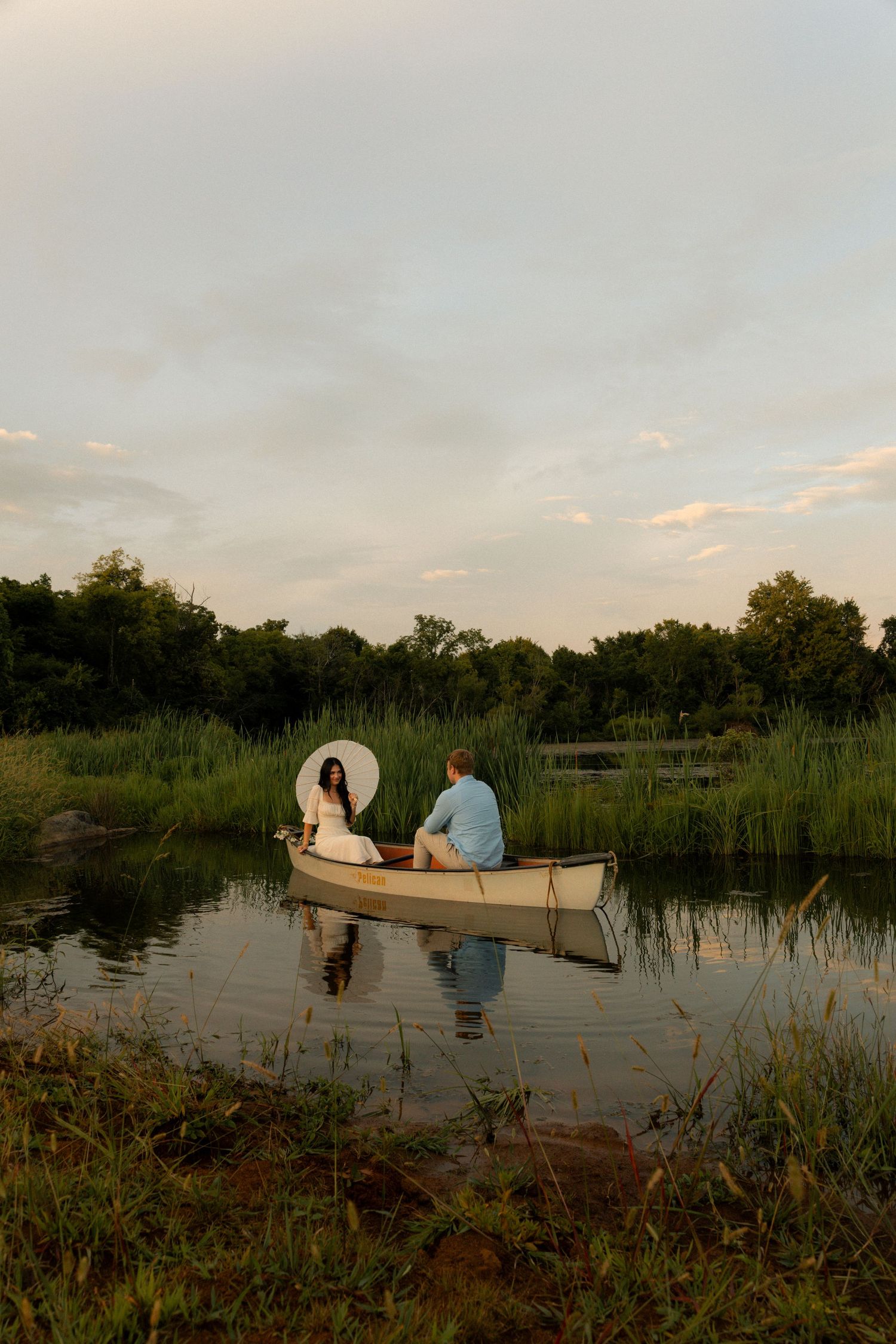 Couple sitting in a white canoe on a calm lake surrounded by lush green trees at sunset.