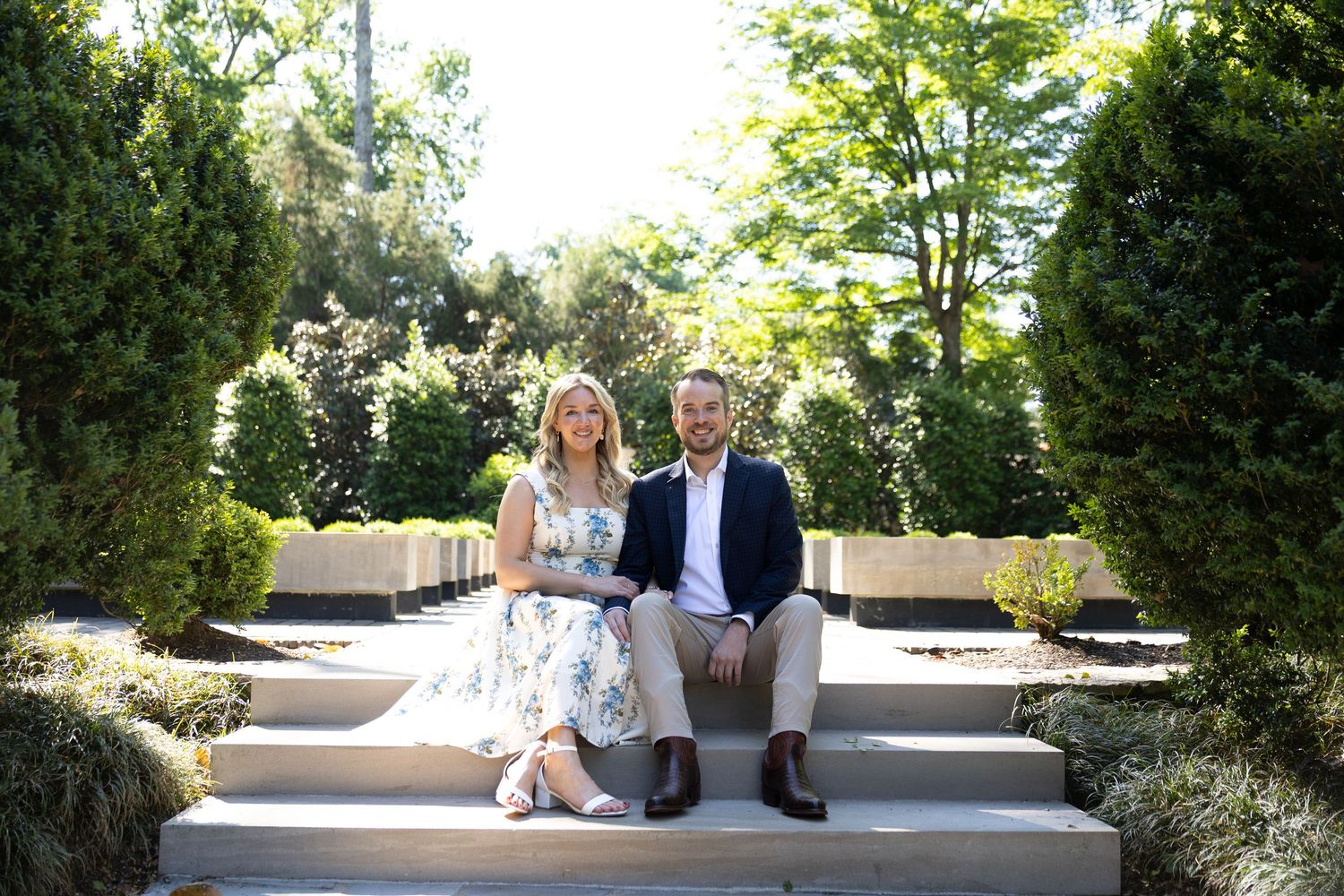 A smiling couple sits together on stone steps surrounded by lush green hedges and trees in a garden setting.