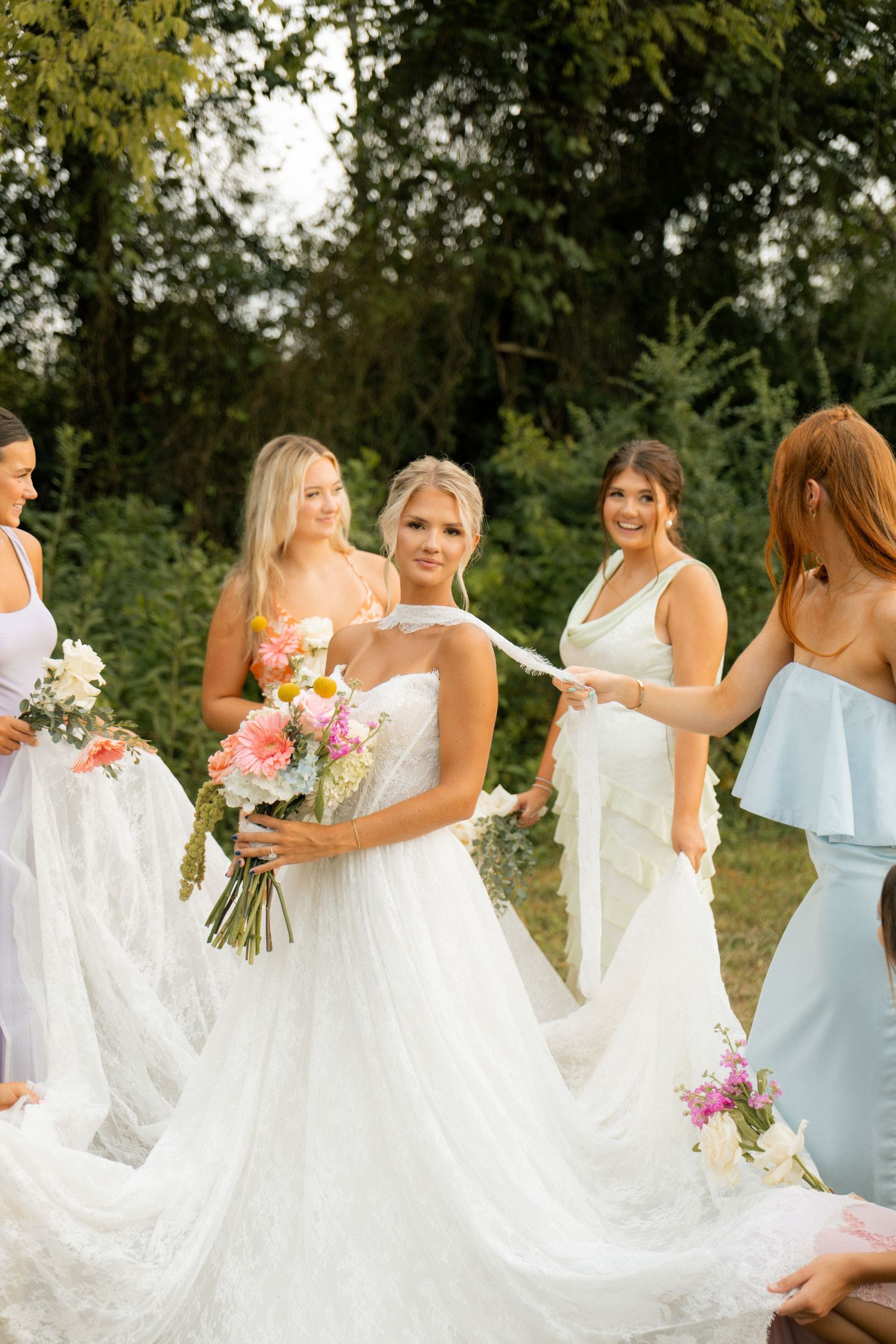 Bride in white strapless gown holds colorful bouquet surrounded by bridesmaids in white and blue dresses outdoors.