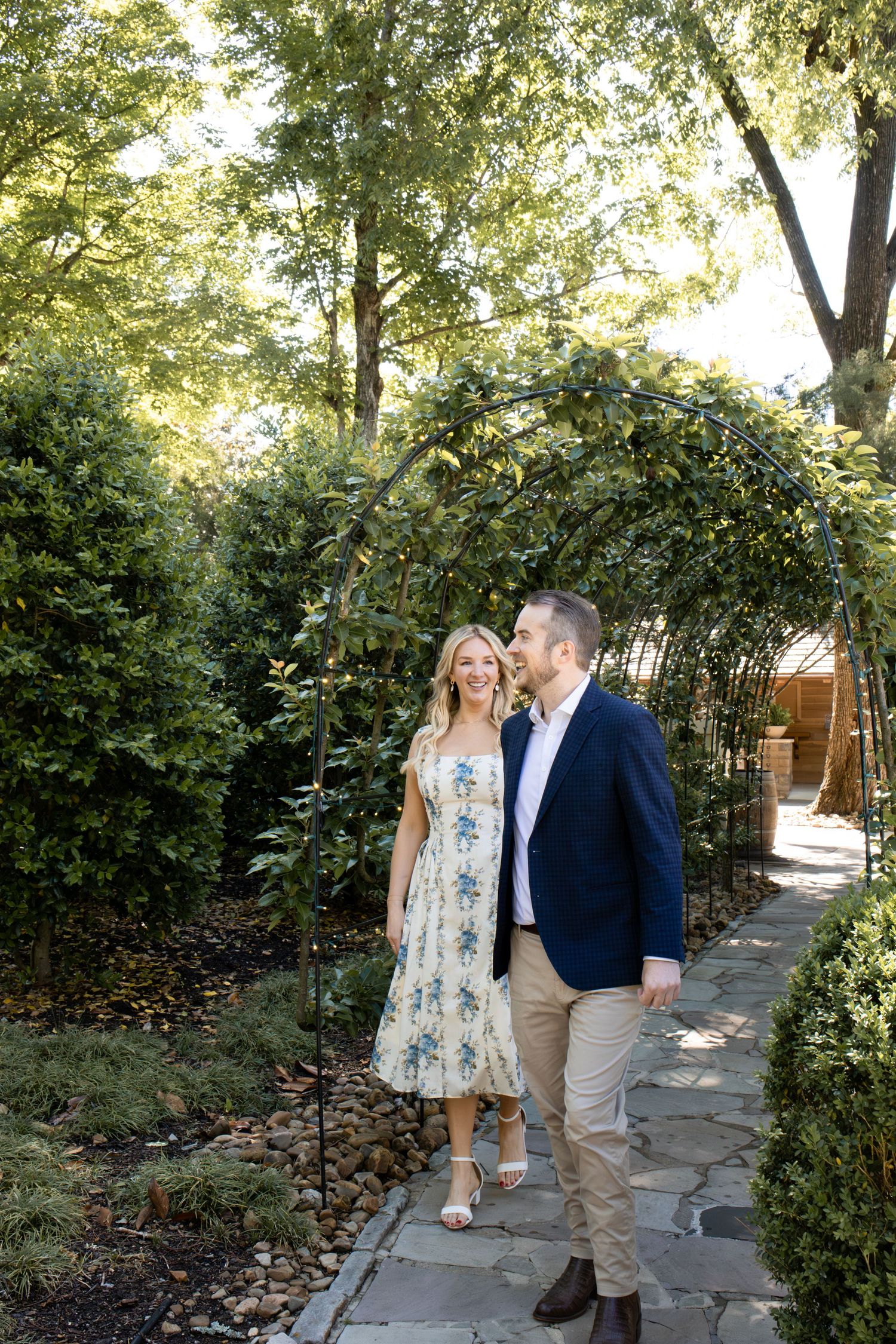 A smiling couple walks hand-in-hand under a metal garden arch surrounded by lush greenery on a stone pathway.