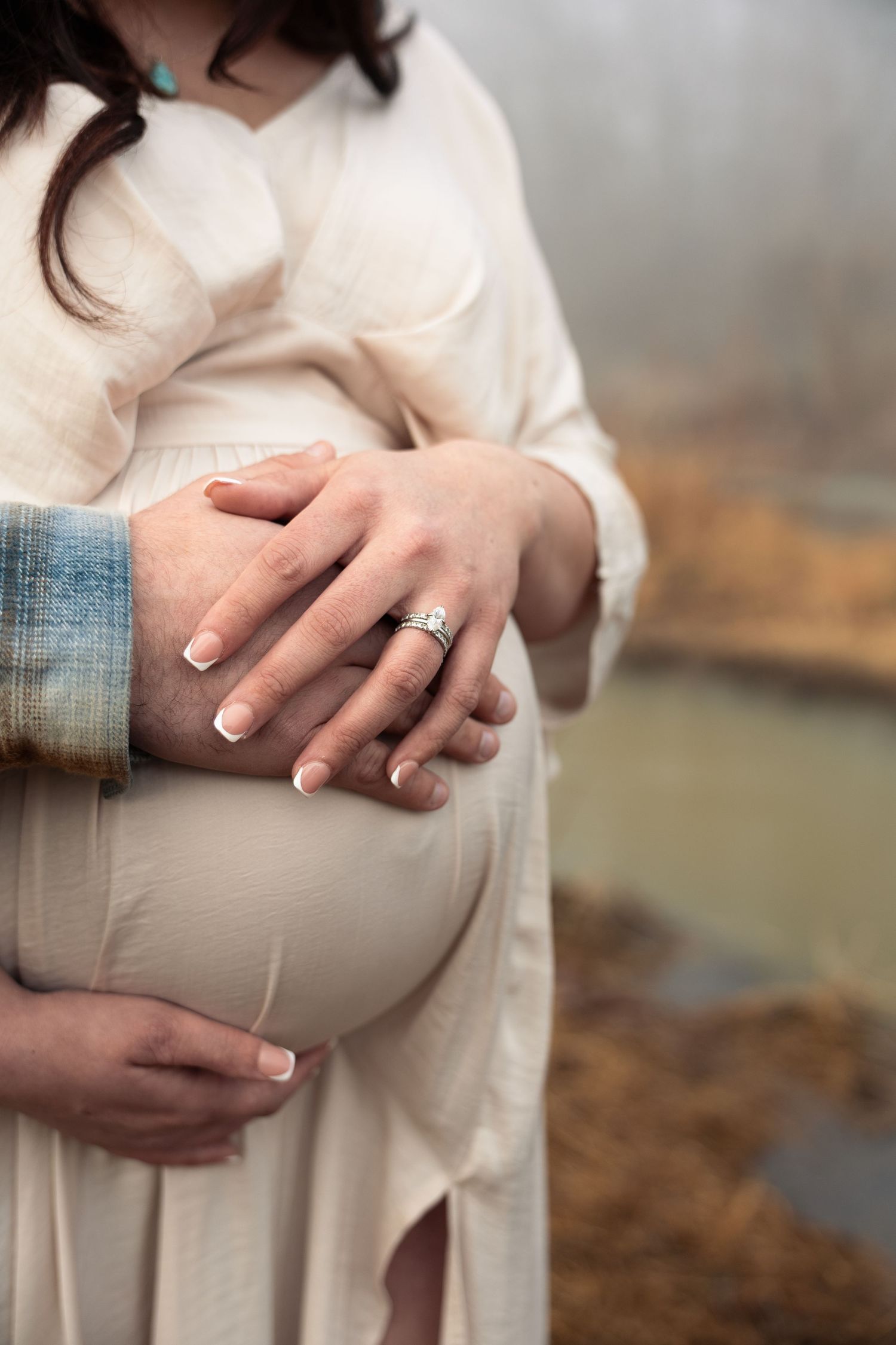 Close-up maternity photo showing hands cradling a pregnant belly in a cream colored dress against a soft blurred background.