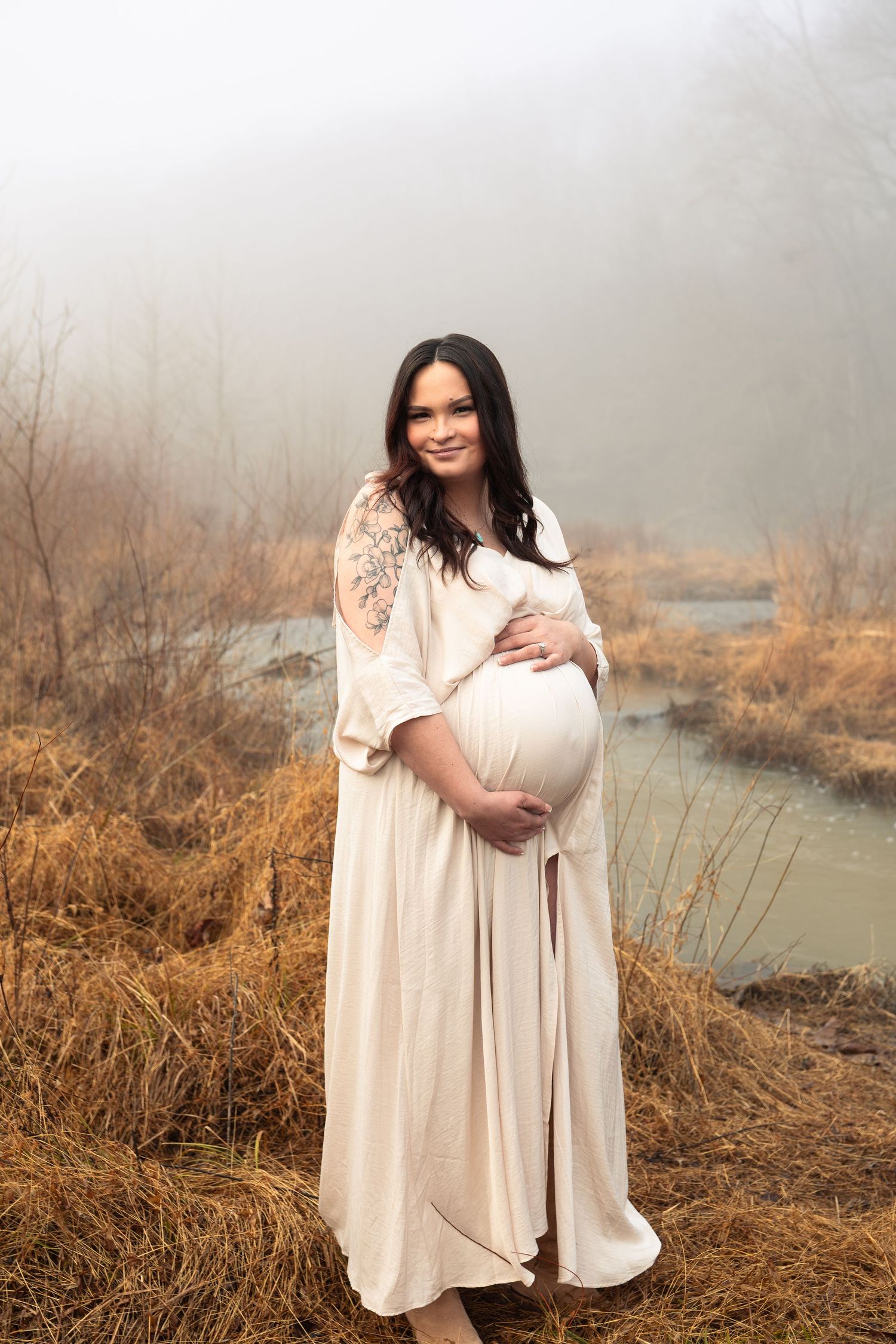 A person in a flowing cream-colored dress stands by a misty river bank surrounded by dried winter grass and brush.
