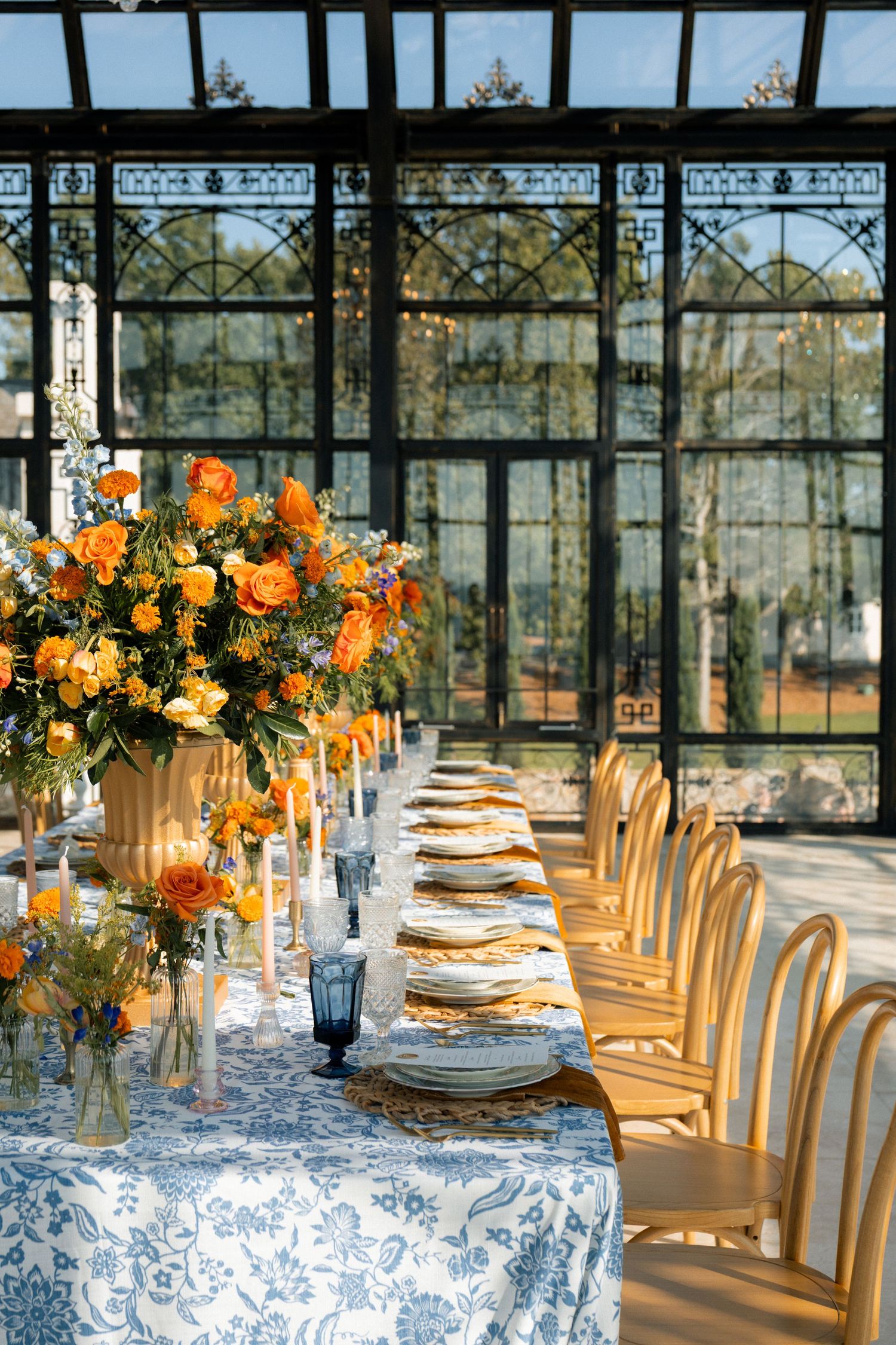 Elegant greenhouse wedding reception with orange floral centerpieces, blue floral tablecloth, gold chargers, and wooden chairs.