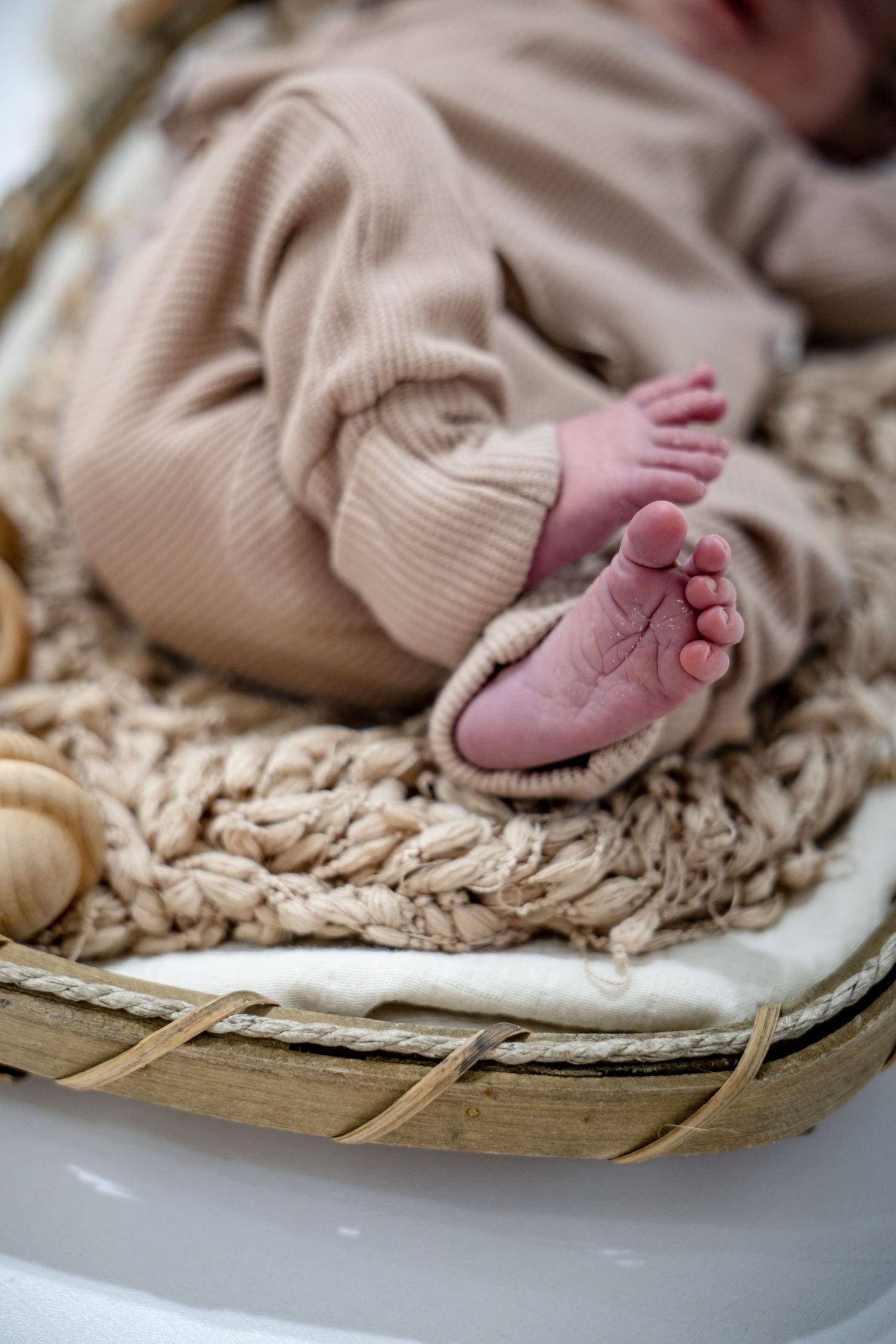 Newborn baby feet peeking out of a beige romper while resting in a woven basket with soft knit blanket.