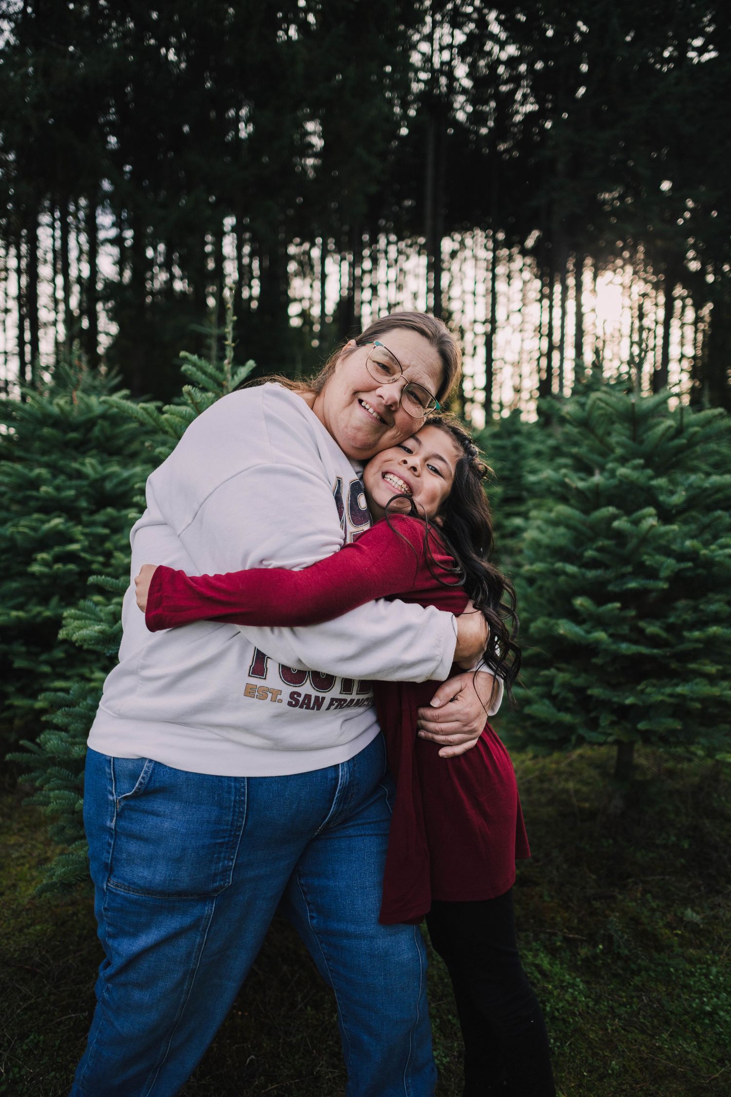 Two people share a warm embrace while standing in front of evergreen trees at dusk.