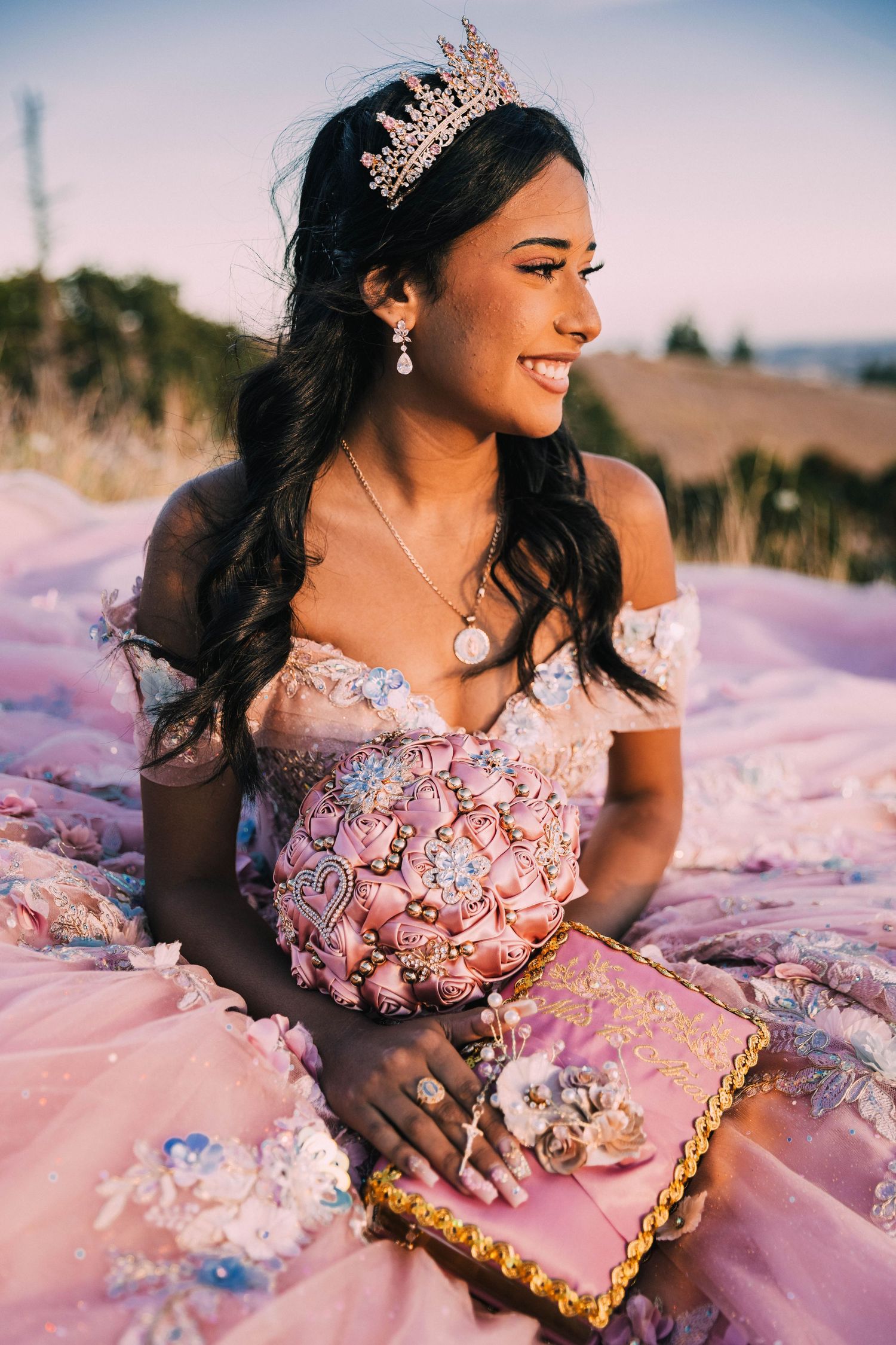A person in a pink quinceanera dress with a tiara poses outdoors during sunset in a dreamy photo session.