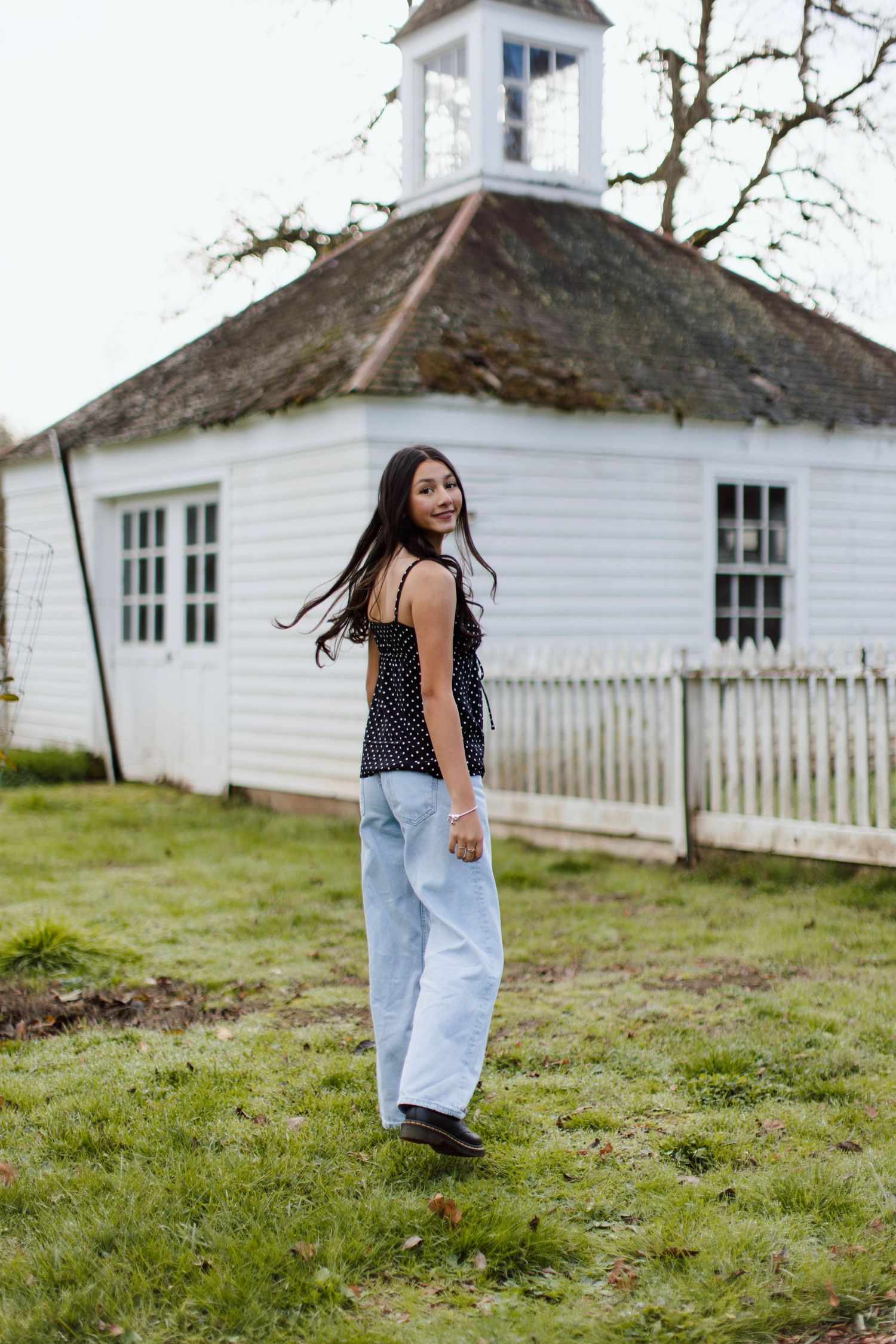 Person in black top and light jeans walks across grassy field near white historic buildings on cloudy day.