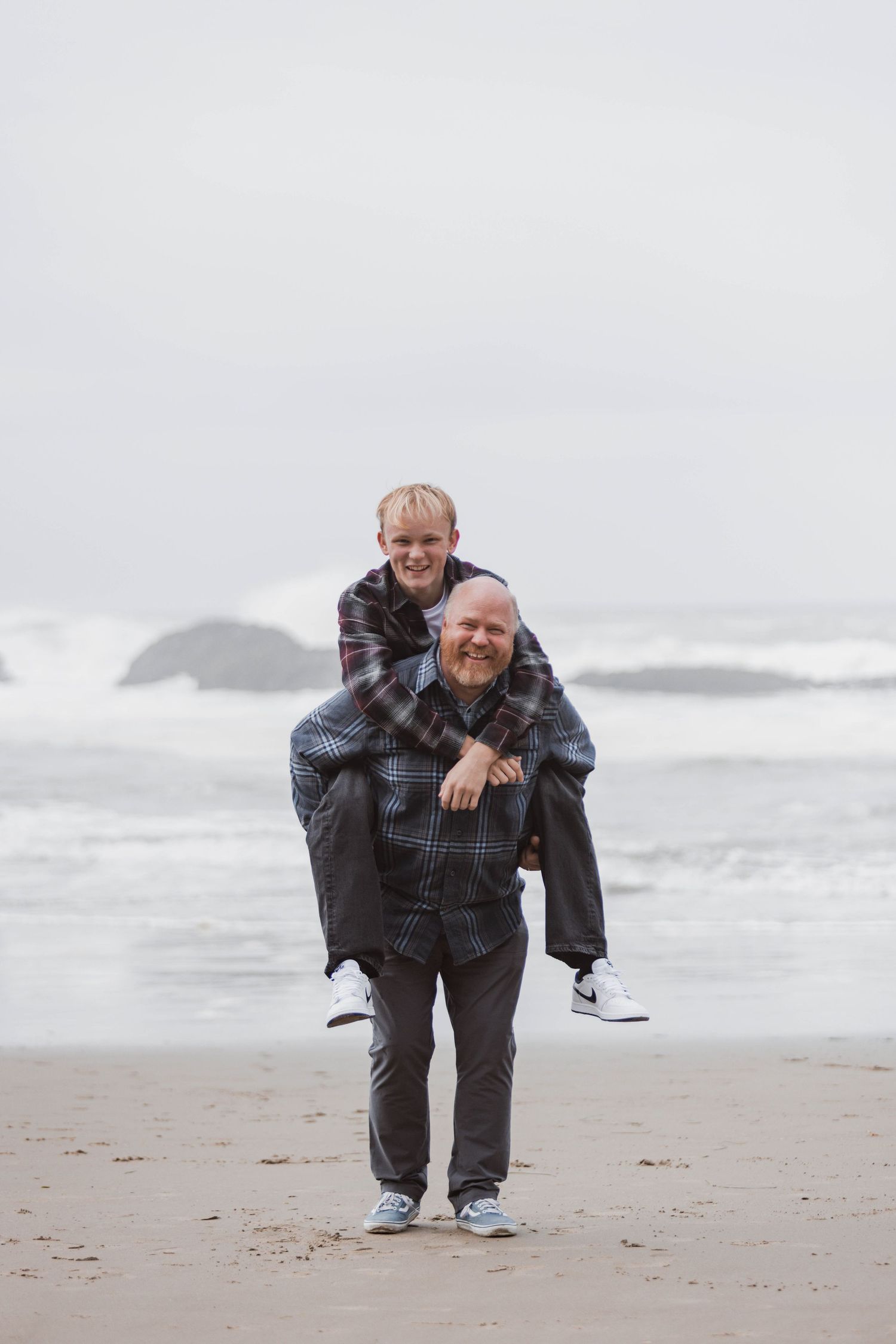 Person in plaid shirt and dark pants crouches on misty beach with waves and rocky coastline in background.