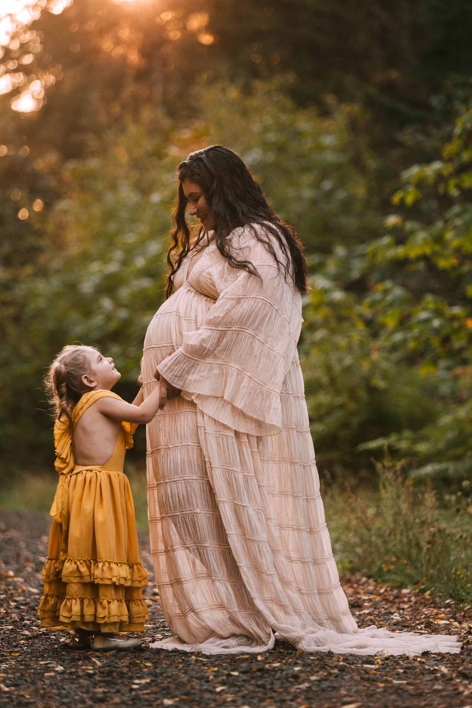 A parent and child in flowing dresses share a tender moment while standing among autumn leaves in a forest setting.