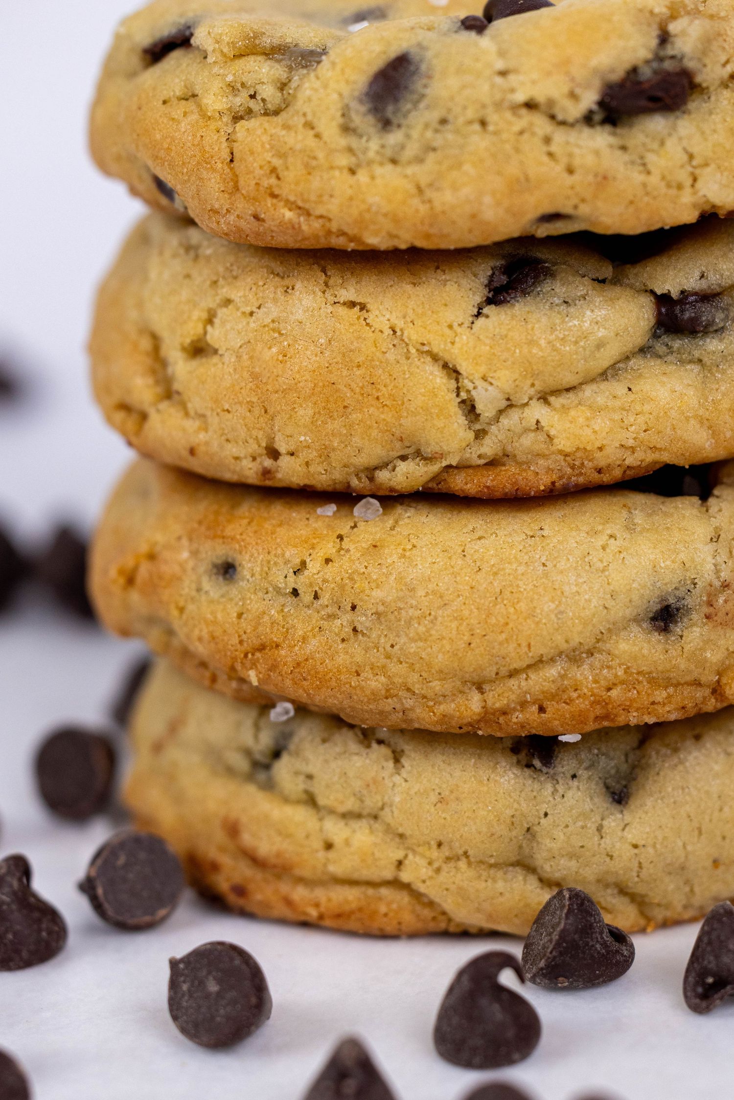 Homemade chocolate chip cookies stacked in rows with scattered chocolate chips on a white surface.