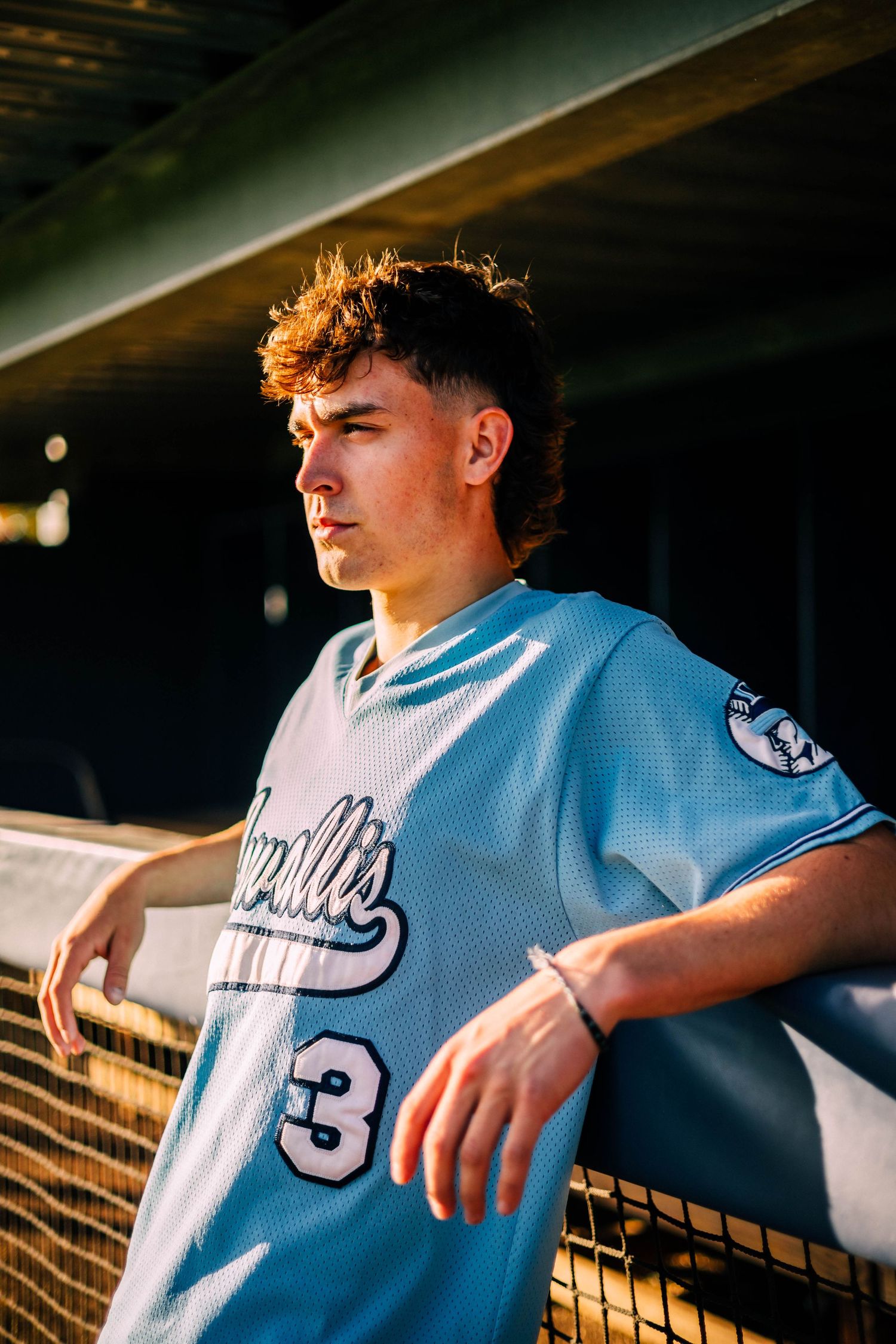 Baseball player in light blue jersey leans against dugout railing during golden hour.