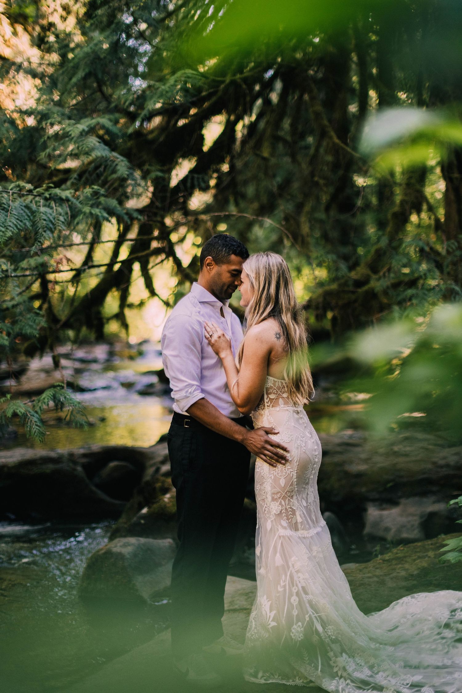 A romantic couple embraces by a serene stream in a lush forest setting, with sunlight filtering through the trees.