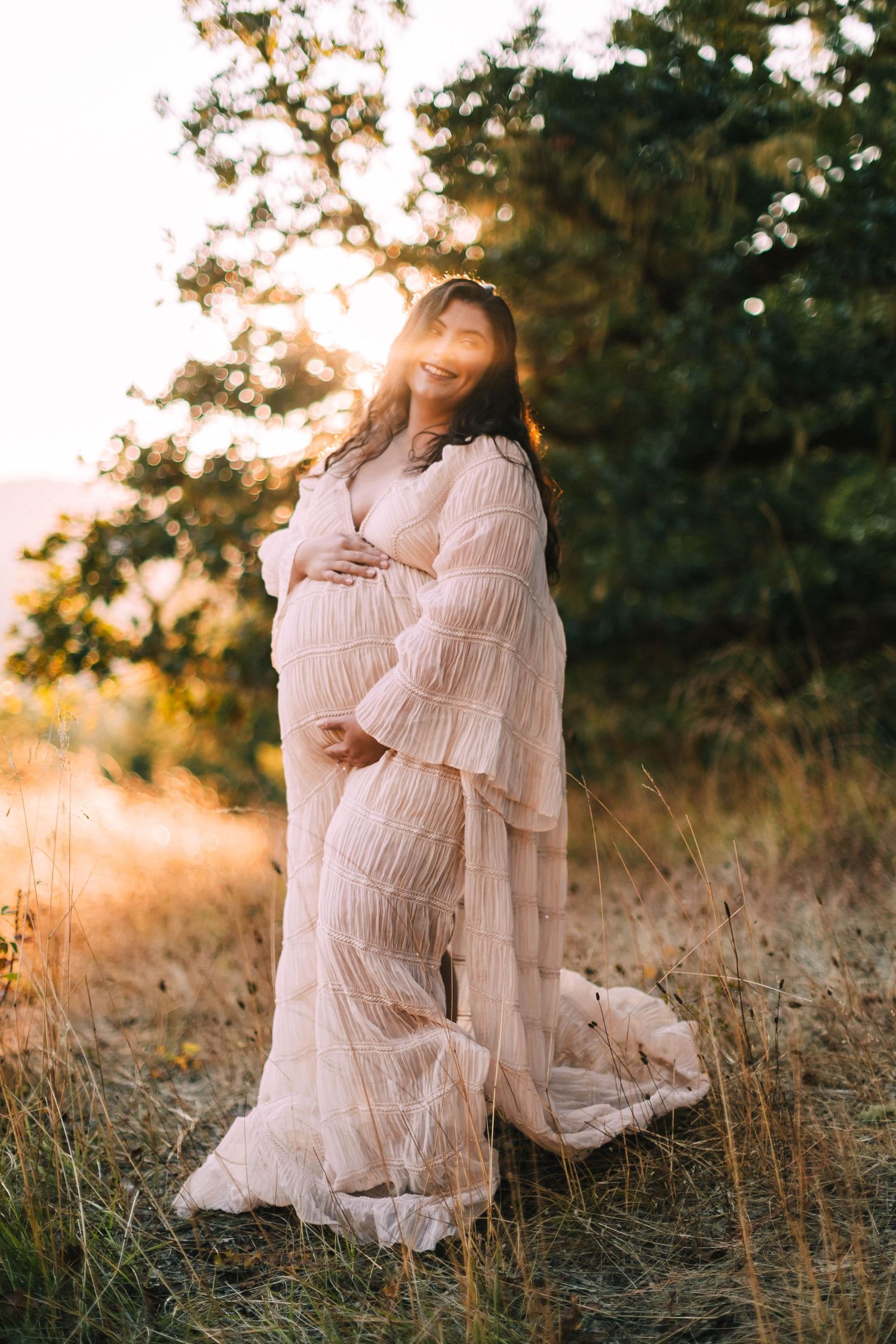 Maternity photoshoot in a golden field at sunset featuring a woman in a flowing pink lace dress.