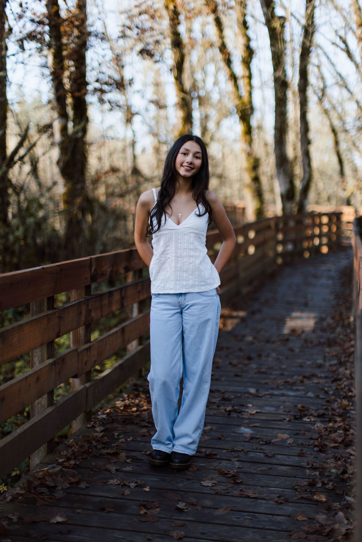 A person in a white top and jeans poses on a wooden boardwalk path through autumn woods at sunset.